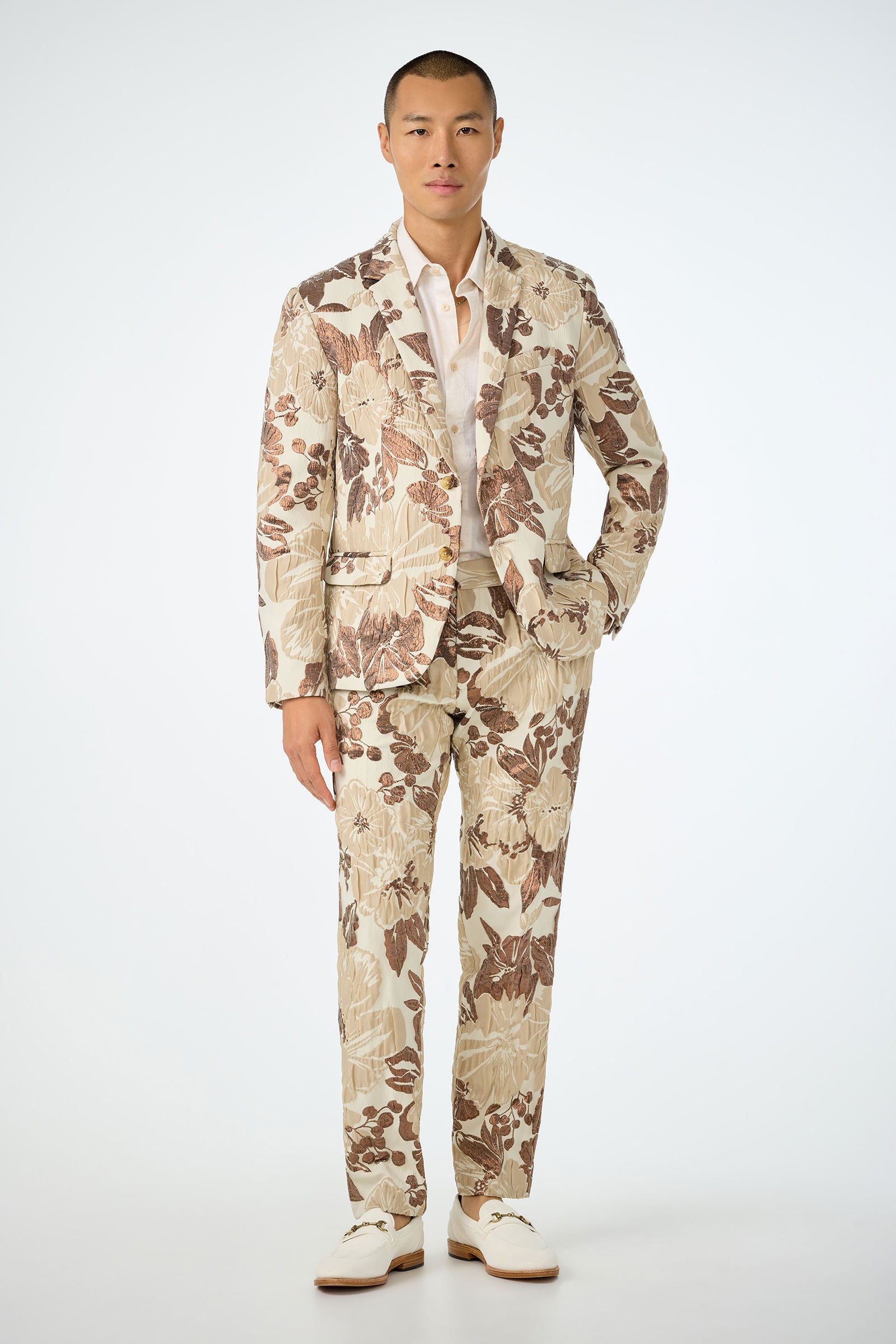 A man with short hair and a neutral expression stands facing forward against a plain white background, wearing the Clyde Slim Trouser in Daintree Jacquard—a cream suit with a bold brown and beige floral pattern—paired with a white shirt and shoes.