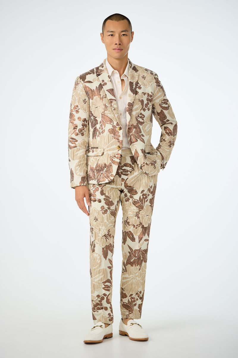 A man with short hair and a neutral expression stands facing forward against a plain white background, wearing the Clyde Slim Trouser in Daintree Jacquard—a cream suit with a bold brown and beige floral pattern—paired with a white shirt and shoes.