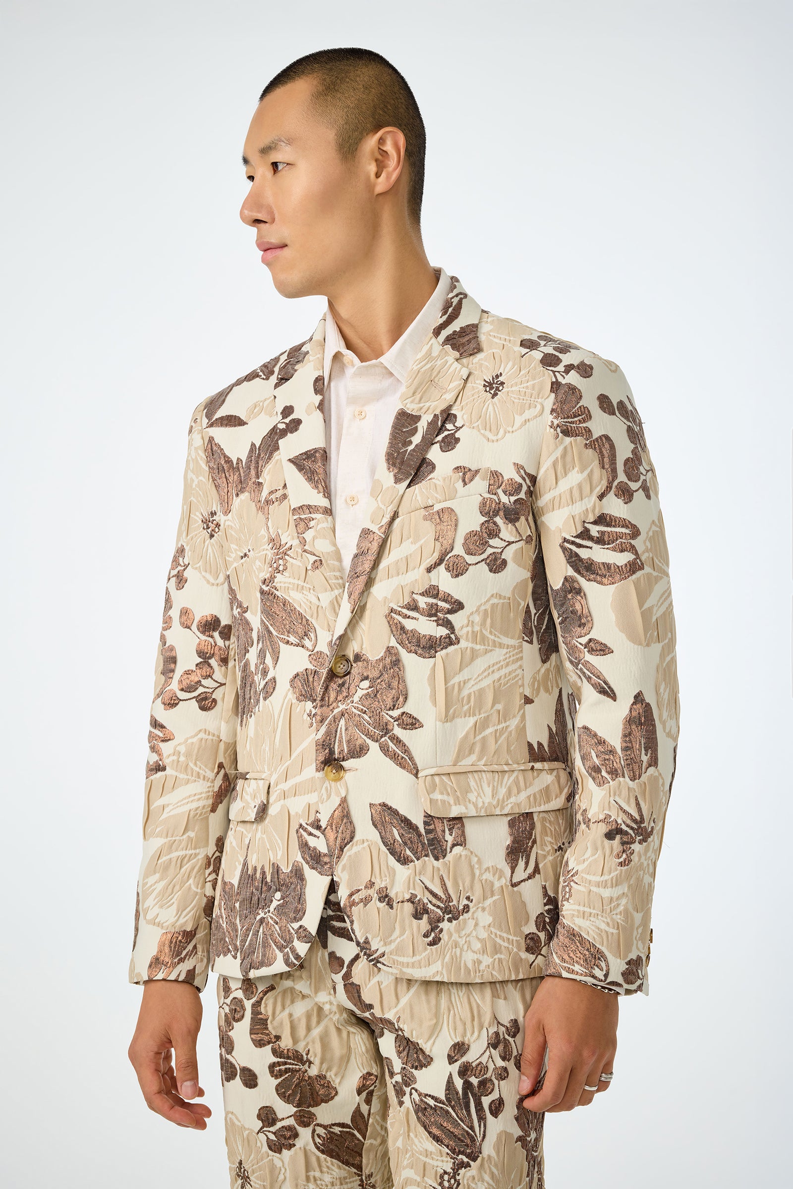 A man with short hair wears the Thurston Blazer - Daintree Jacquard, a cream suit with a bold brown and beige floral pattern, over a light shirt, standing against a plain background and looking to his left.
