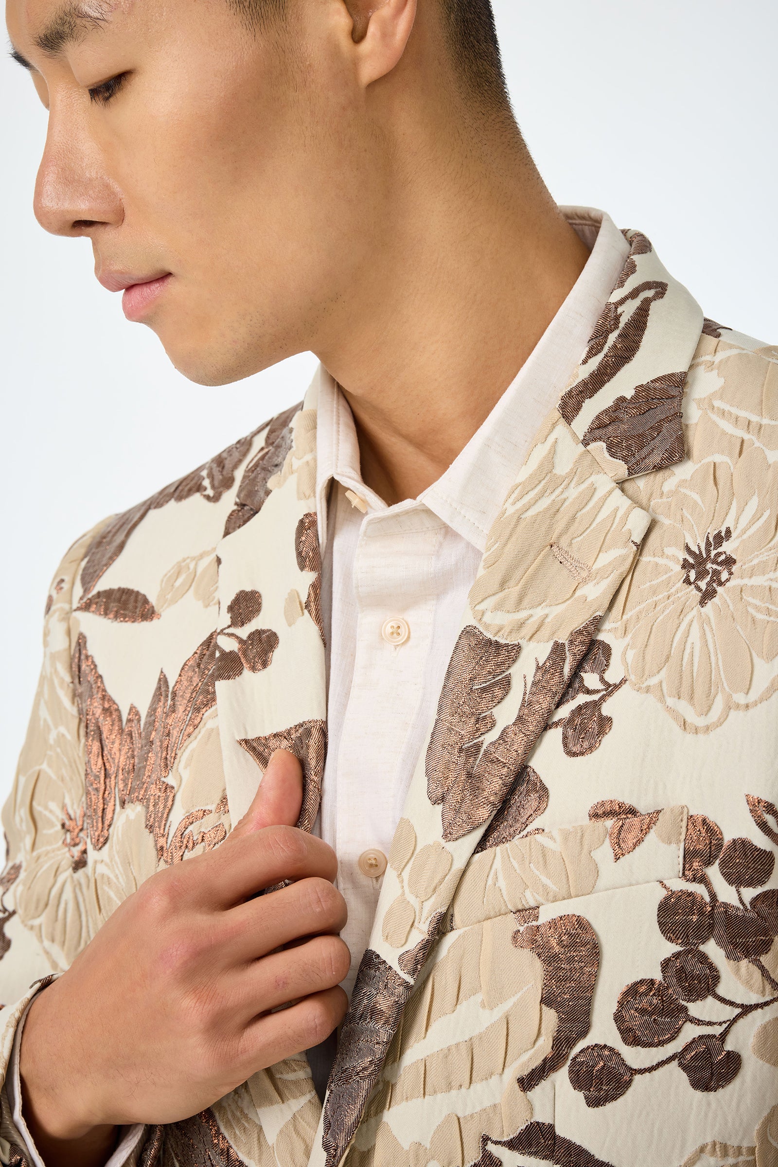 A person in the Thurston Blazer - Daintree Jacquard, featuring a cream base with brown floral patterns, wears it over a light beige button-up and holds the blazers lapel; their face is partially visible in profile.