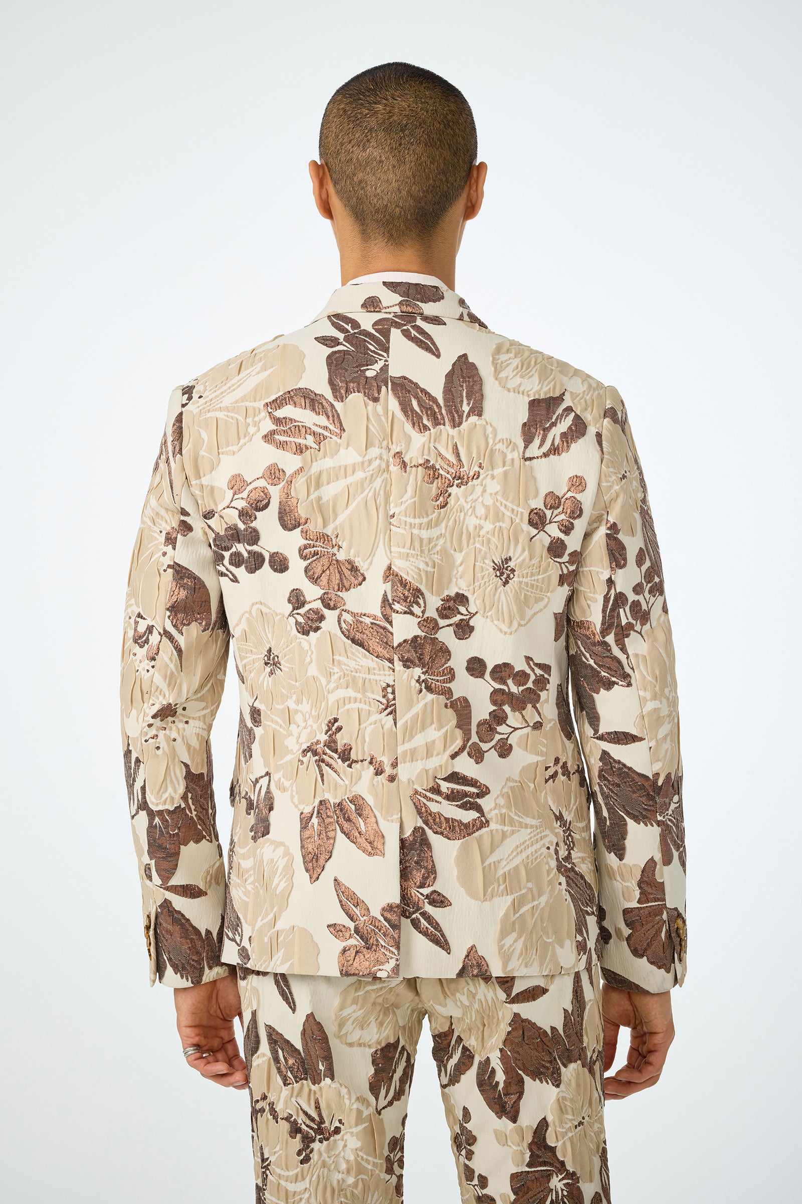 A person with short hair is seen from behind wearing the Thurston Blazer - Daintree Jacquard, a cream-colored suit featuring a bold brown and beige floral pattern. The plain, light background highlights the vibrant blazer design.
