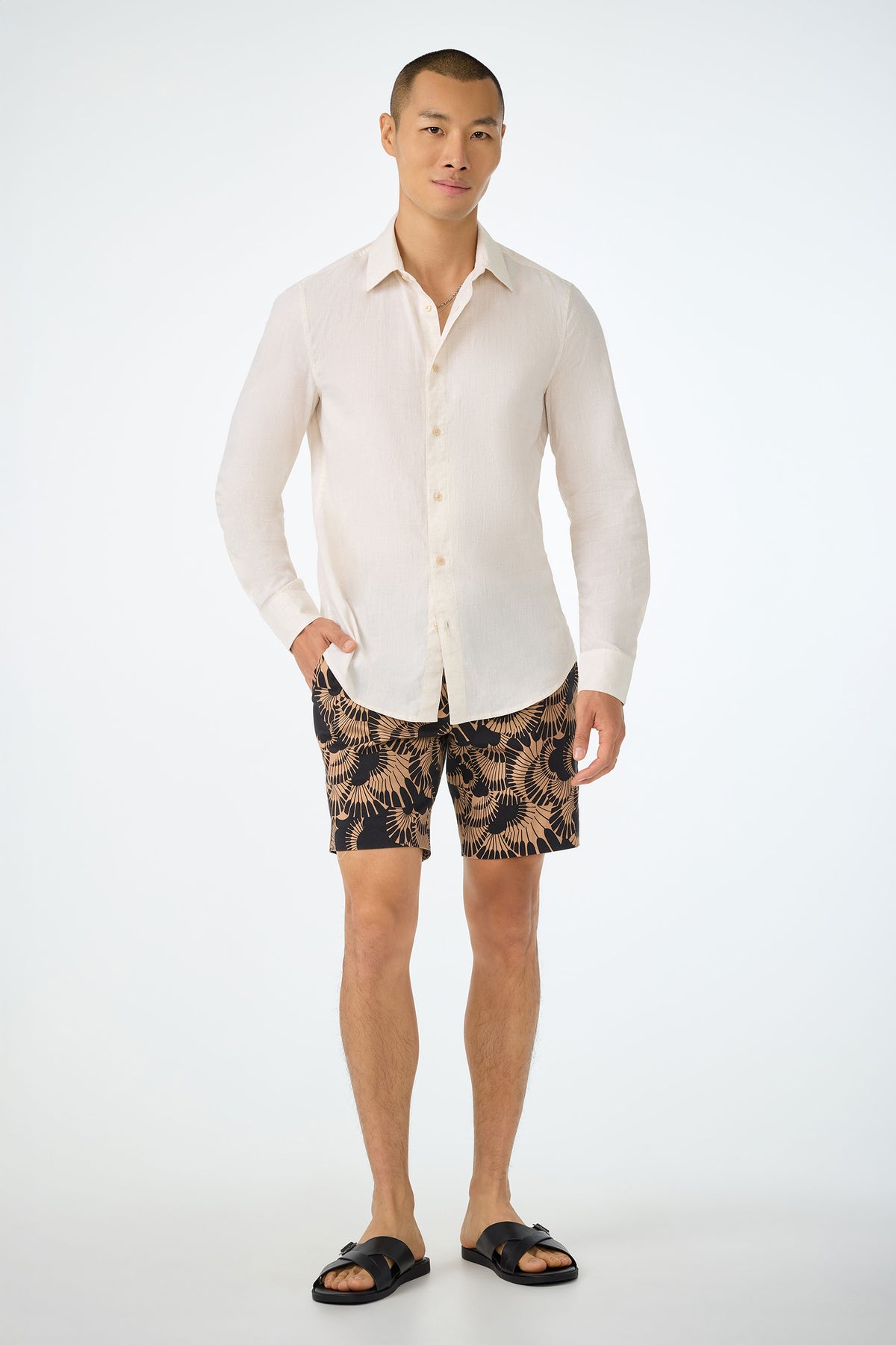 A person stands against a plain background wearing a cream button-up shirt, Lawrence Short - Juniper Herringbone, and black slide sandals. They face forward with one hand in their pocket, smiling slightly.
