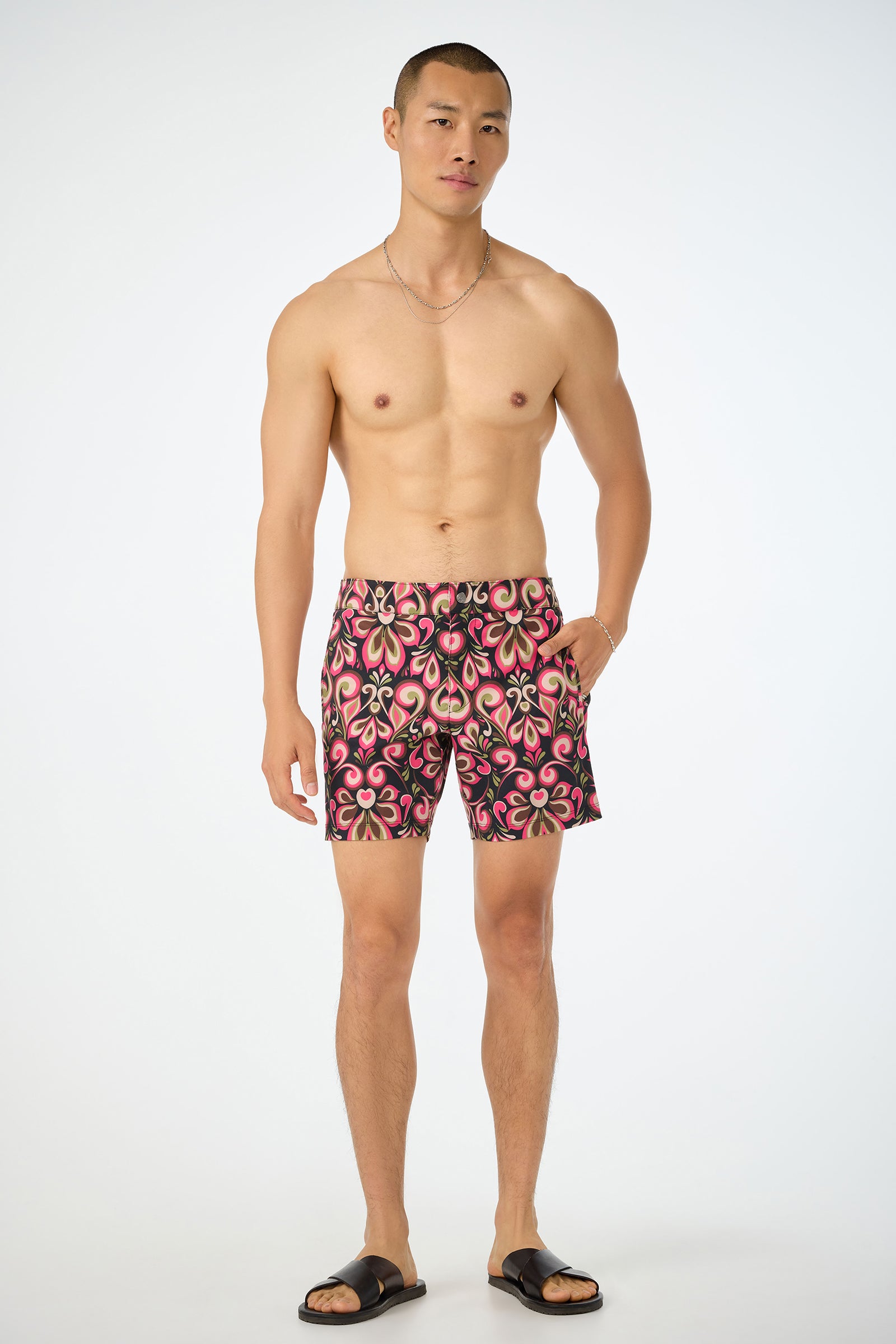 Wearing the Honolulu Board Short in the Sanctuary print, a man poses against a white background with one hand in his pocket, black slide sandals, and a thin chain necklace. The shorts feature pink, black, and beige patterns.