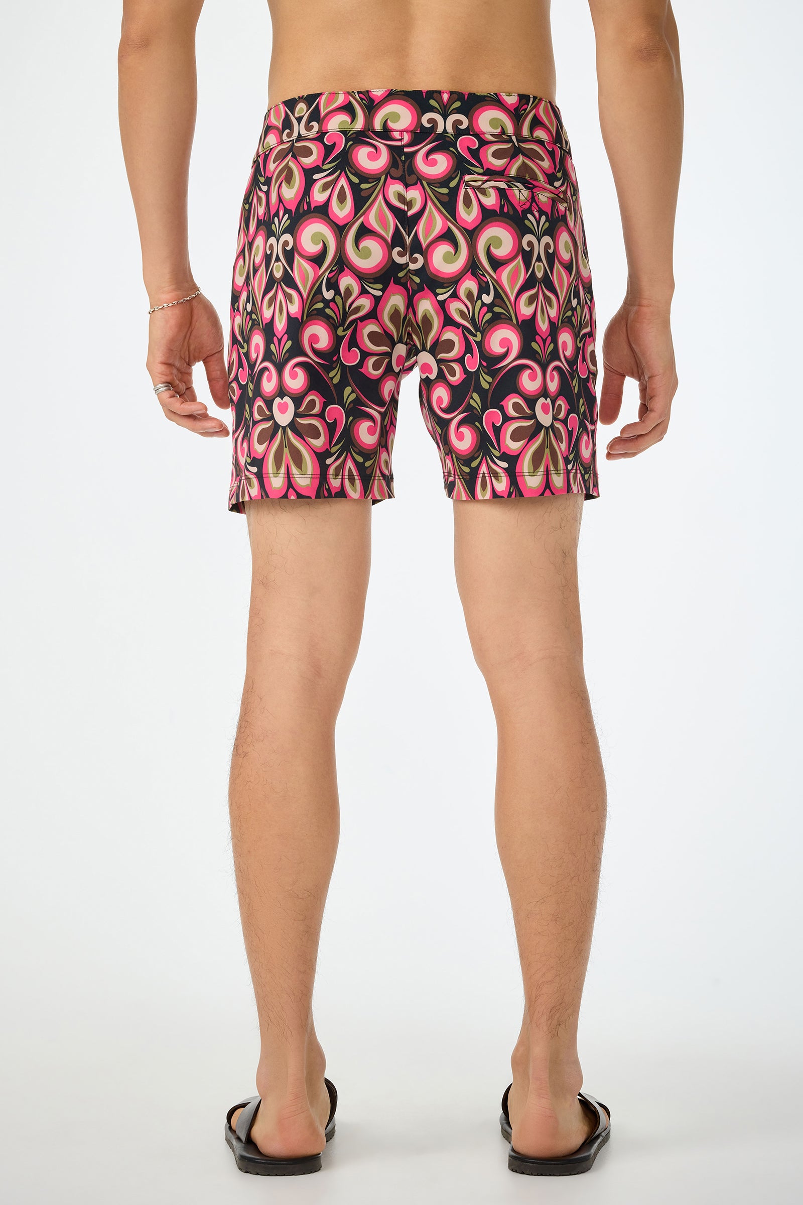 A person faces away from the camera, wearing black flip-flops and the Honolulu Board Short - Sanctuary, featuring a bold, colorful retro swirl pattern in pink, red, cream, and black. The background is plain white.