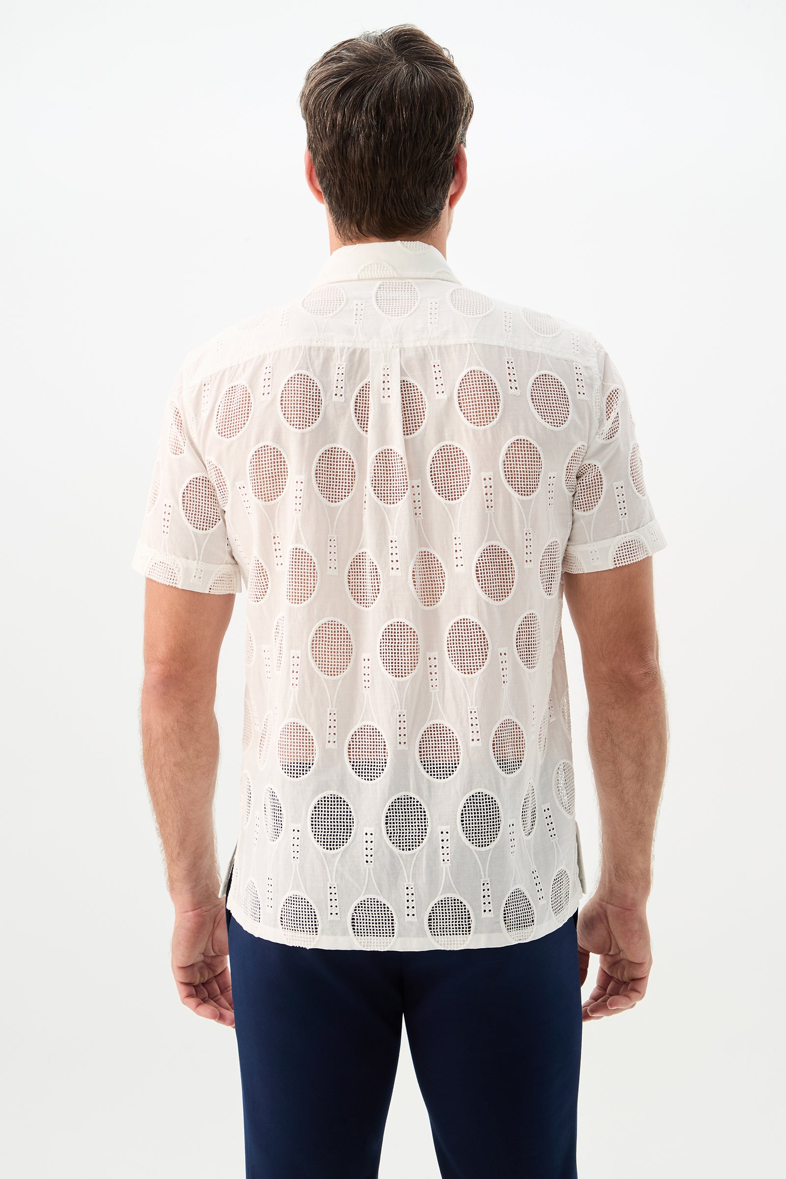 A man is shown from the back wearing the Branson Shirt - Tennis Eyelet, a short-sleeve, white, semi-sheer shirt with a circular geometric pattern, paired with dark pants against a plain white background.