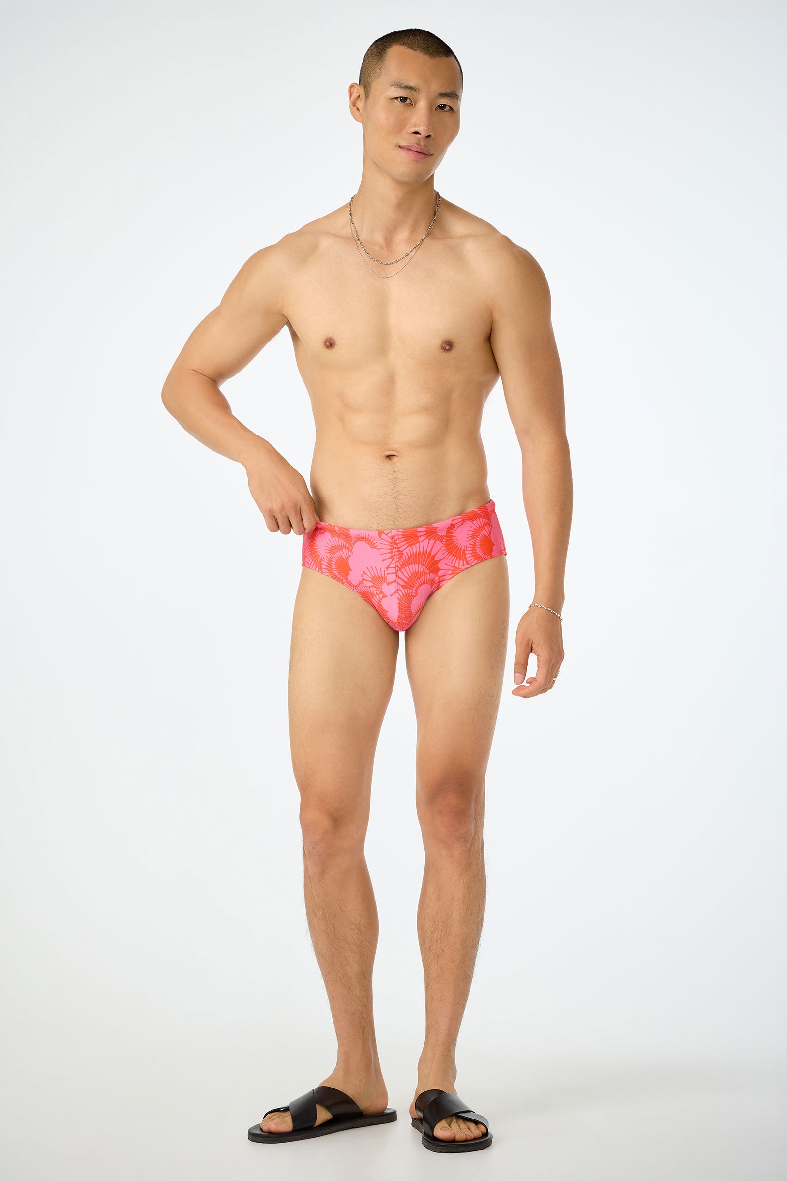 A man stands against a plain background wearing the San Gregorio Swim Brief - Juniper, black sandals, and a necklace, adjusting the side of his swimwear.