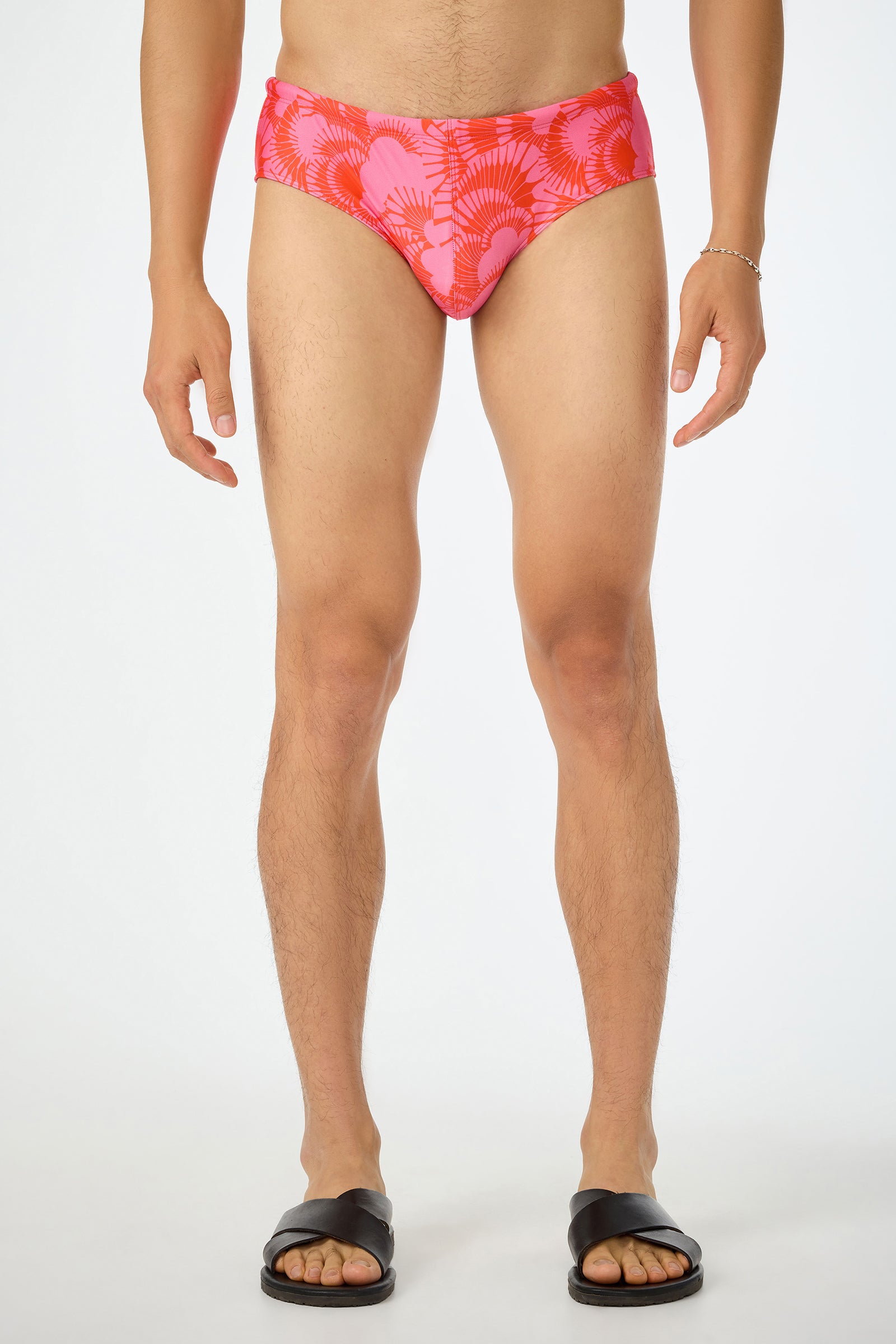 A person stands facing forward, wearing the San Gregorio Swim Brief - Juniper (Multi), black slide sandals, and a silver bracelet. The background is plain white, and the persons head is not visible in the image.