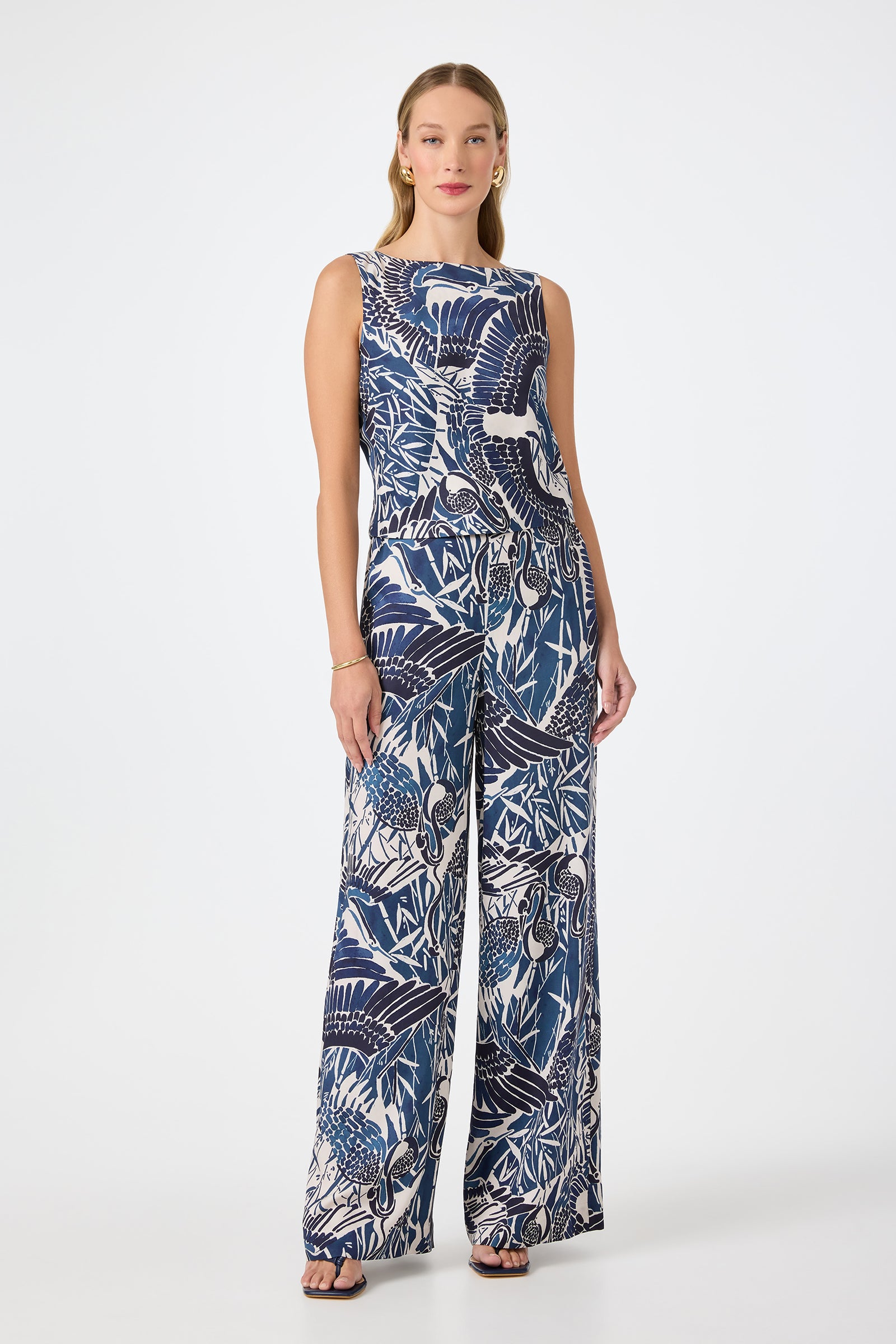 A woman with long, wavy hair and a neutral expression stands against a white background wearing the Hakuto Tie-Back Top - Tsuru, which features a sleeveless design and bold blue and white tropical pattern, paired with black sandals.