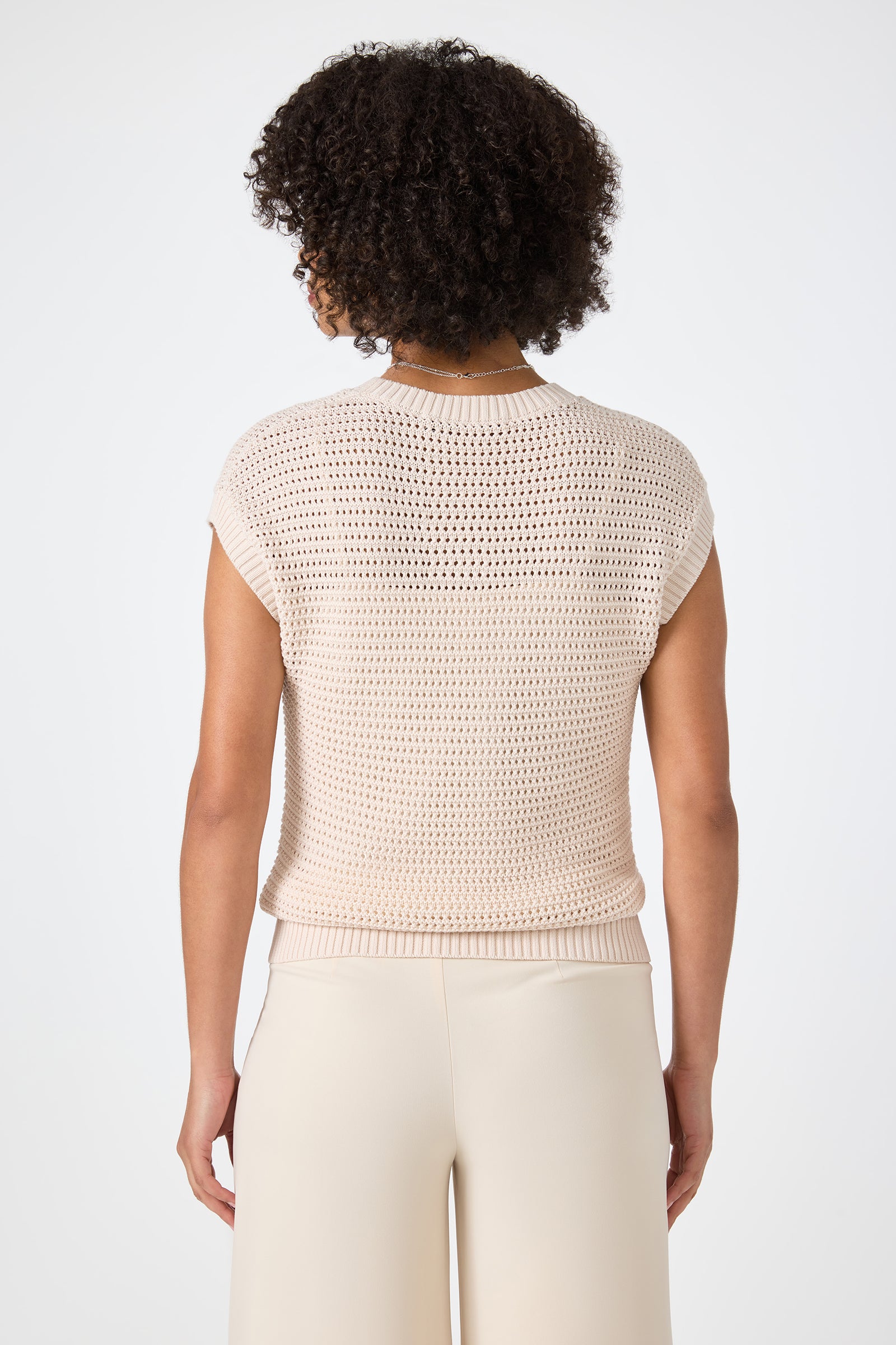 A person with short, curly hair is shown from the back, wearing the Yuri Knit Pullover in light beige and cream-colored pants against a plain white background.
