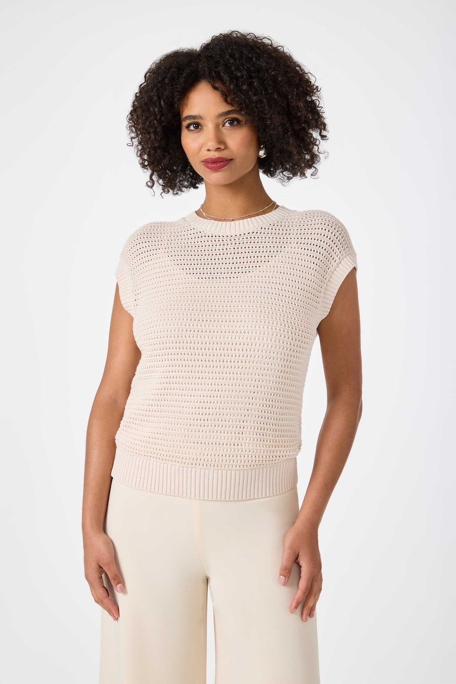 A woman with curly hair wears the Yuri Knit Pullover in Sancerre, featuring short sleeves, as she stands against a plain white background and looks at the camera with a slight smile.
