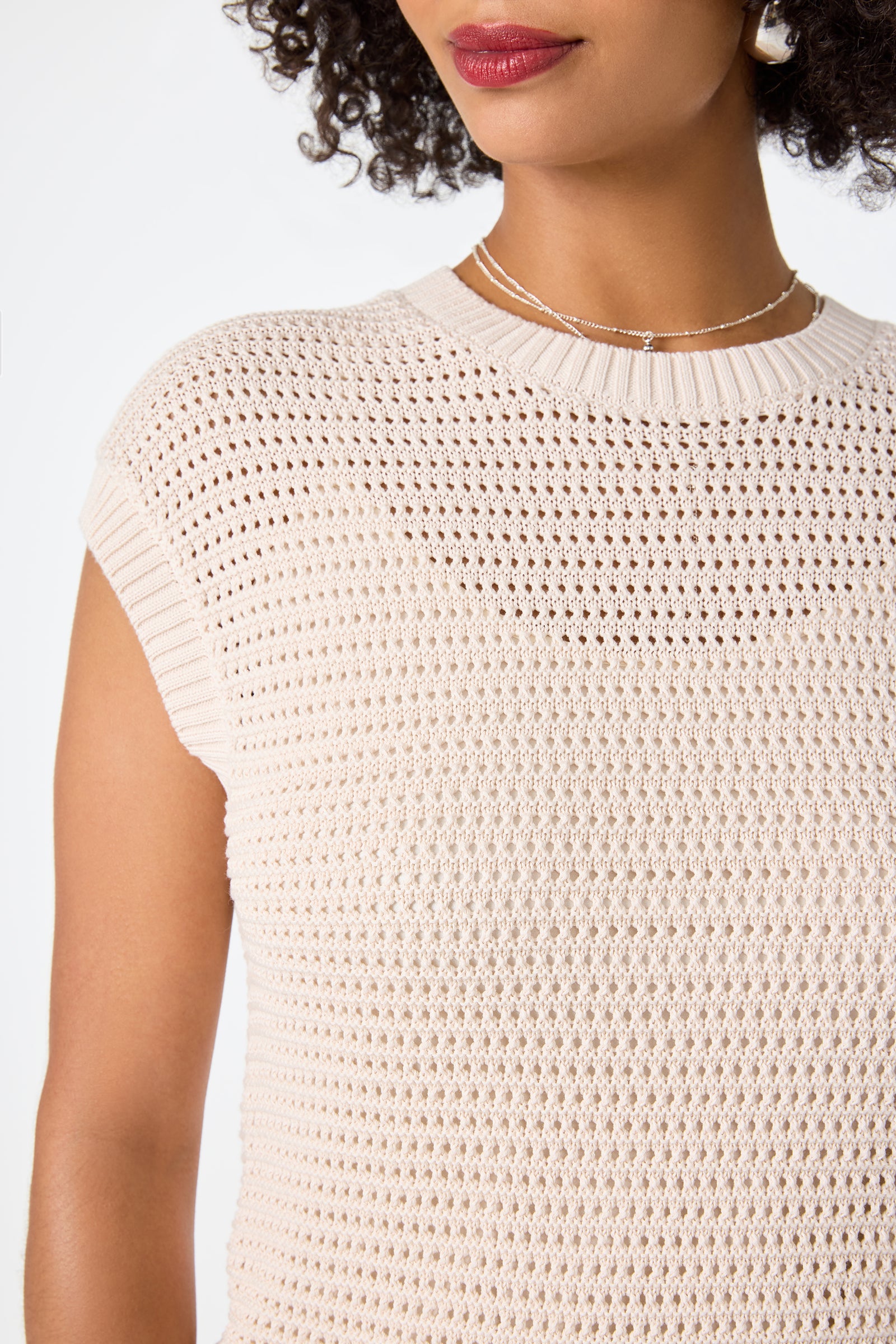 A close-up of a woman with curly hair and red lipstick wearing the Yuri Knit Pullover—a cream-colored, short-sleeved knit top with a textured pattern, styled with a delicate necklace and earring.