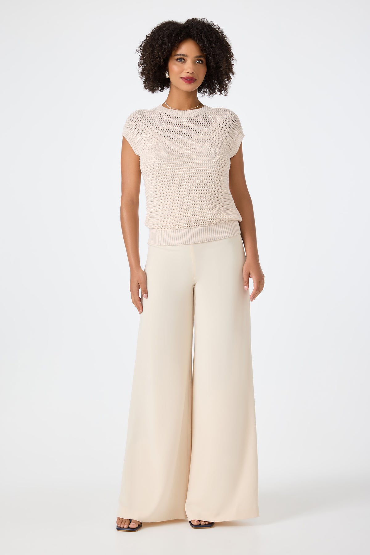 A woman with curly hair wears the Yuri Knit Pullover, a short-sleeve, light beige top, paired with wide-leg, high-waisted cream pants against a plain white background.