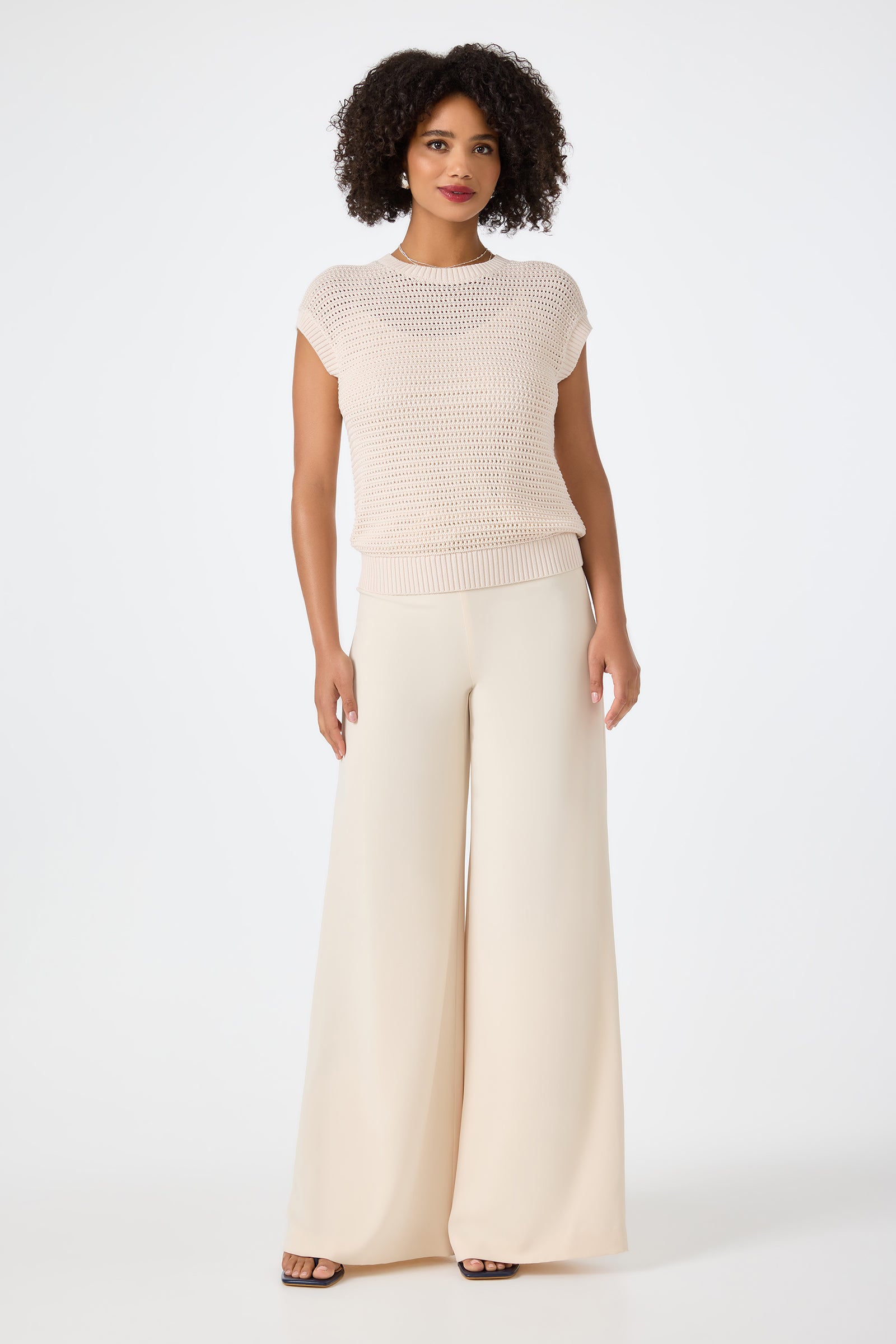 A woman with curly hair wears the Yuri Knit Pullover, a short-sleeve, light beige top, paired with wide-leg, high-waisted cream pants against a plain white background.