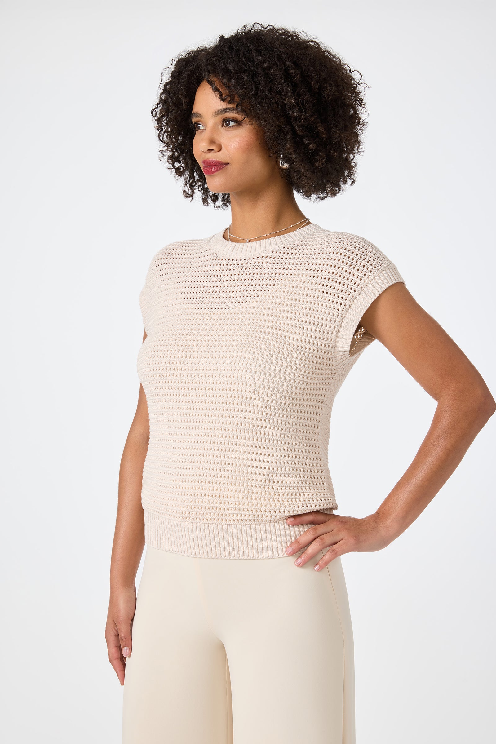 A woman with curly hair wears the Yuri Knit Pullover, a light beige short-sleeve top, paired with cream pants, standing with one hand on her hip and looking sideways against a plain white background.