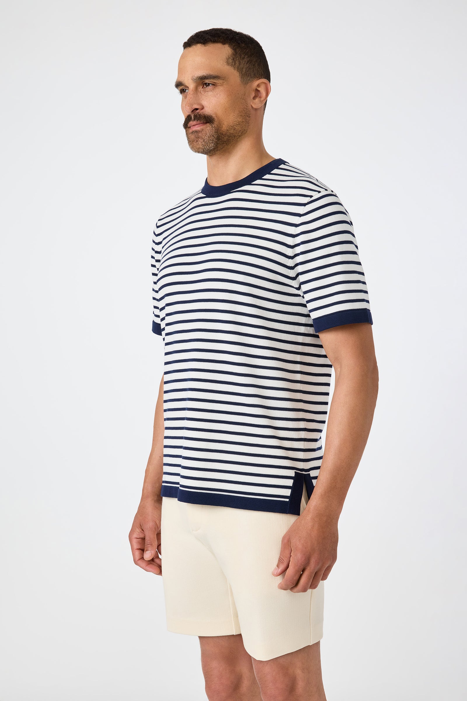 A man with short hair and a beard wears the Giovanni Stripe Short-Sleeve Pullover in white and navy blue stripes with cream-colored shorts, standing against a plain light background.