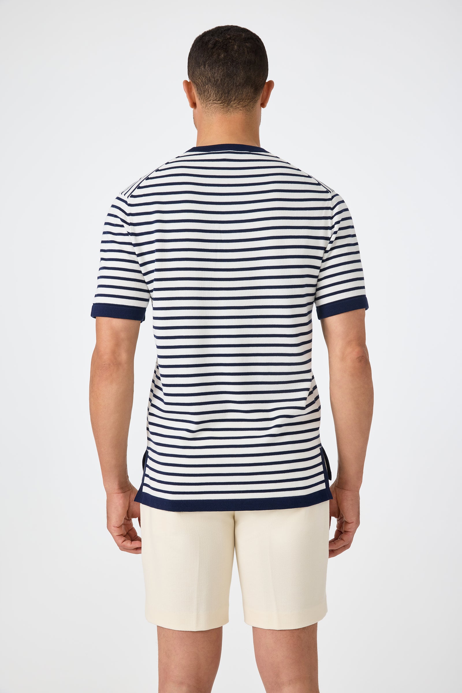 A man faces away from the camera, wearing the Giovanni Stripe Short-Sleeve Pullover—white with navy horizontal stripes and trim—paired with light shorts. The background is plain and light.