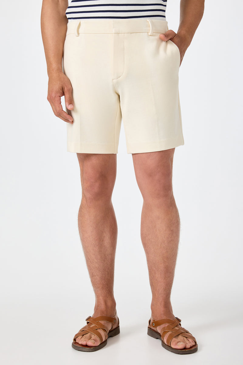 Wearing the Lawrence Short in ecru, paired with a navy and white striped shirt and brown sandals, a person stands with one hand in their pocket against a plain white background.
