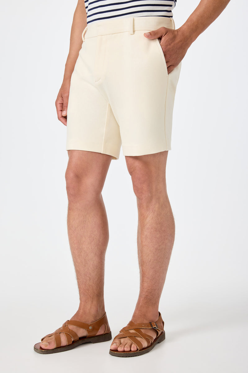 A person wearing the Lawrence Short in Ecru stands with one hand in their pocket against a plain white background, styled with a white and navy striped shirt and brown sandals.