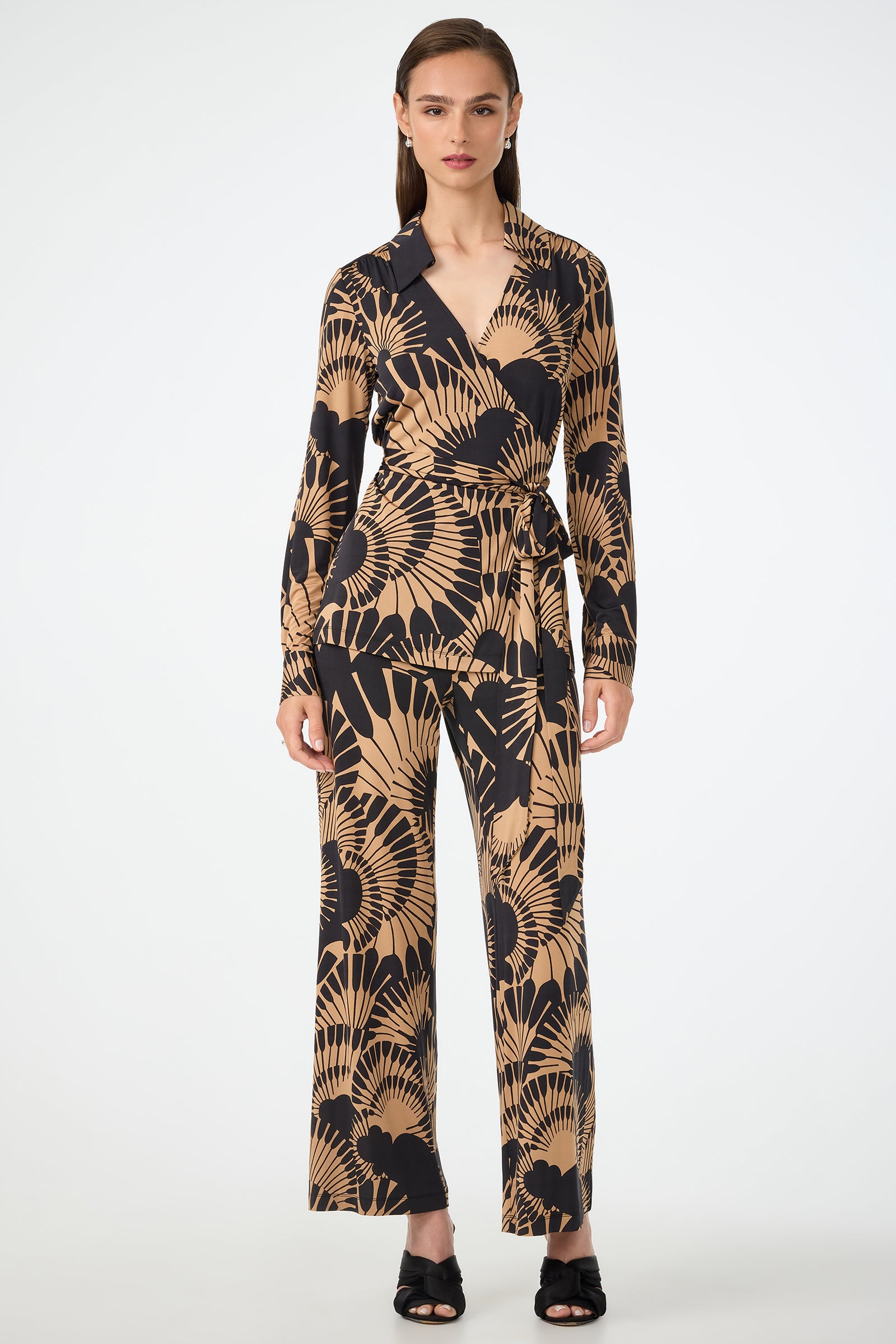 A woman faces forward, wearing the Makeba Wrap Top in Juniper Jersey— a long-sleeve beige and black wrap top with an abstract leaf pattern—paired with matching wide-leg pants. She has straight brown hair and black open-toe heels.