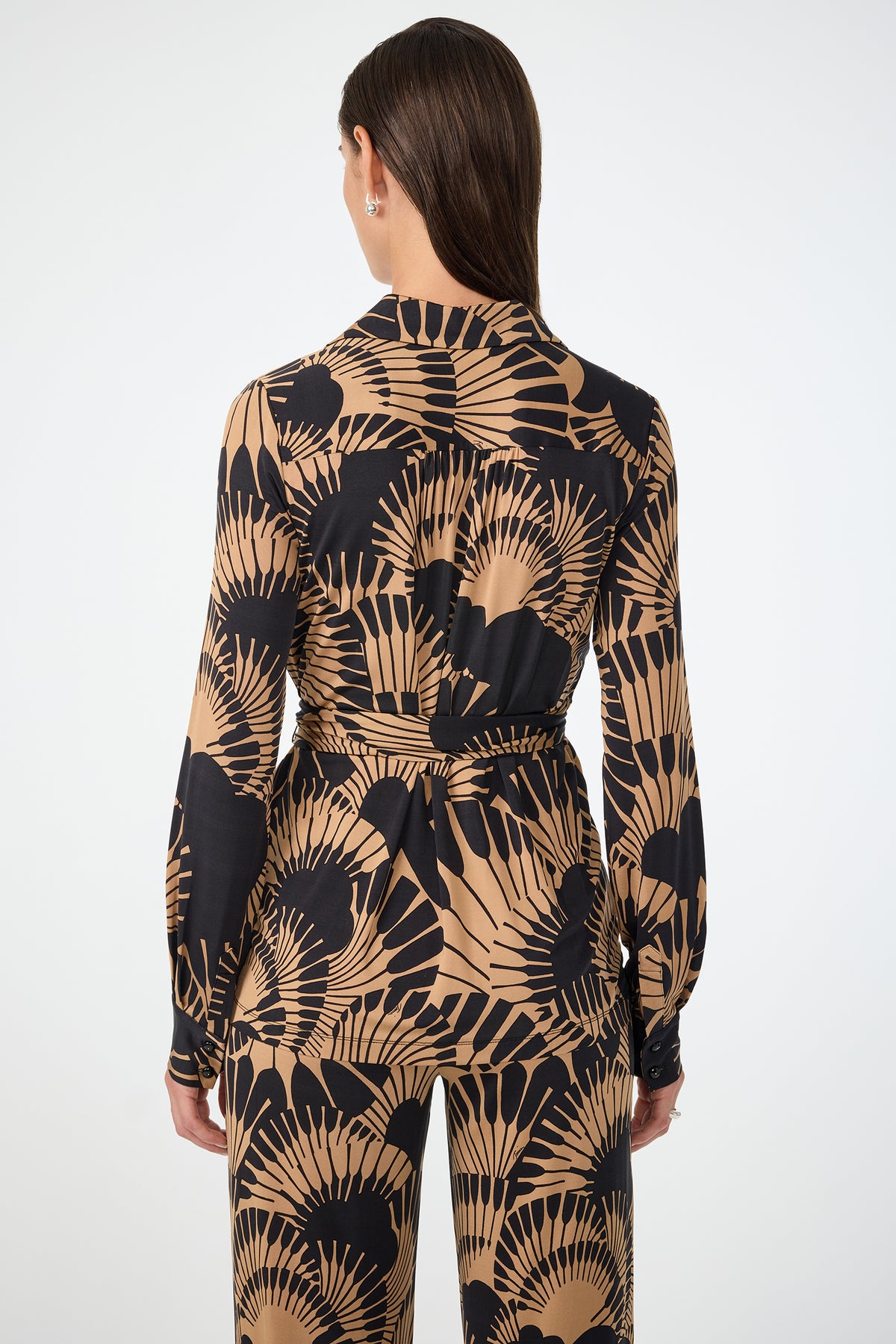 A woman with straight brown hair stands facing away, wearing the Makeba Wrap Top in Juniper Jersey. The top features a tan and black fan pattern, a waist tie, and is styled with matching pants against a plain white background.