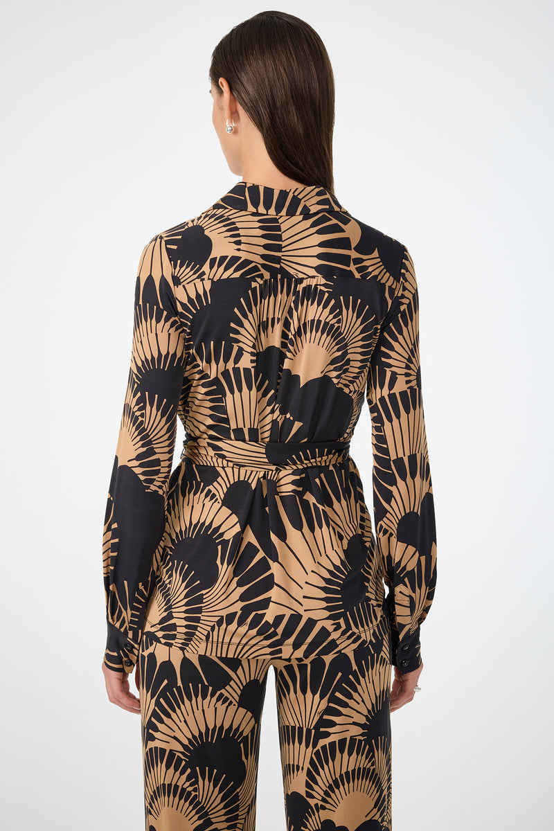 A woman with straight brown hair stands facing away, wearing the Makeba Wrap Top in Juniper Jersey. The top features a tan and black fan pattern, a waist tie, and is styled with matching pants against a plain white background.