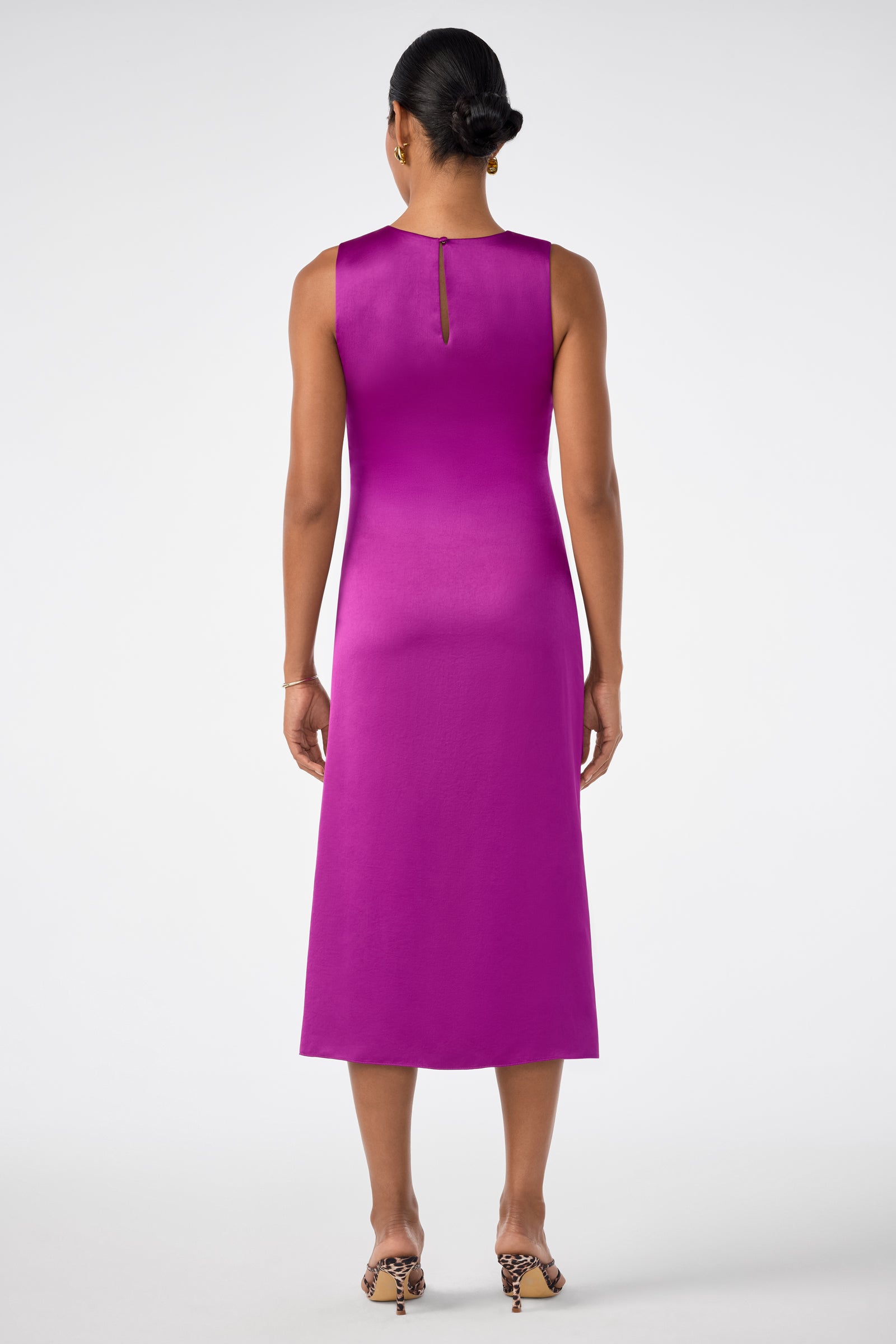 A woman stands facing away, wearing the sleeveless Chestnut Dress in bright magenta with a small keyhole closure at the back and open-toed heeled sandals. Her hair is styled in a low bun.