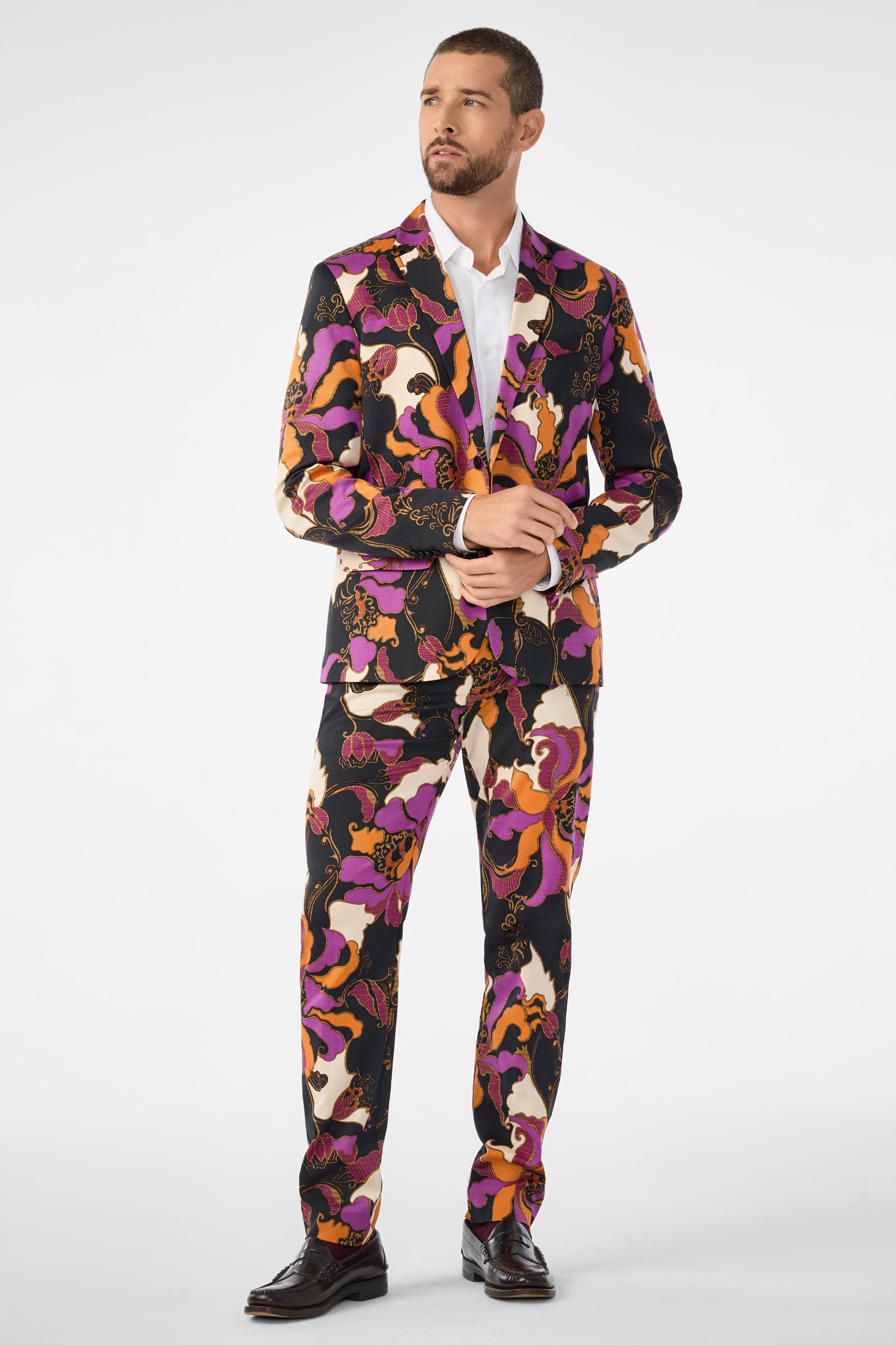 A man models the Clyde Slim Trouser, featuring an abstract pattern in purple, orange, black, and white tones, styled with a white dress shirt and dark shoes against a plain white background.