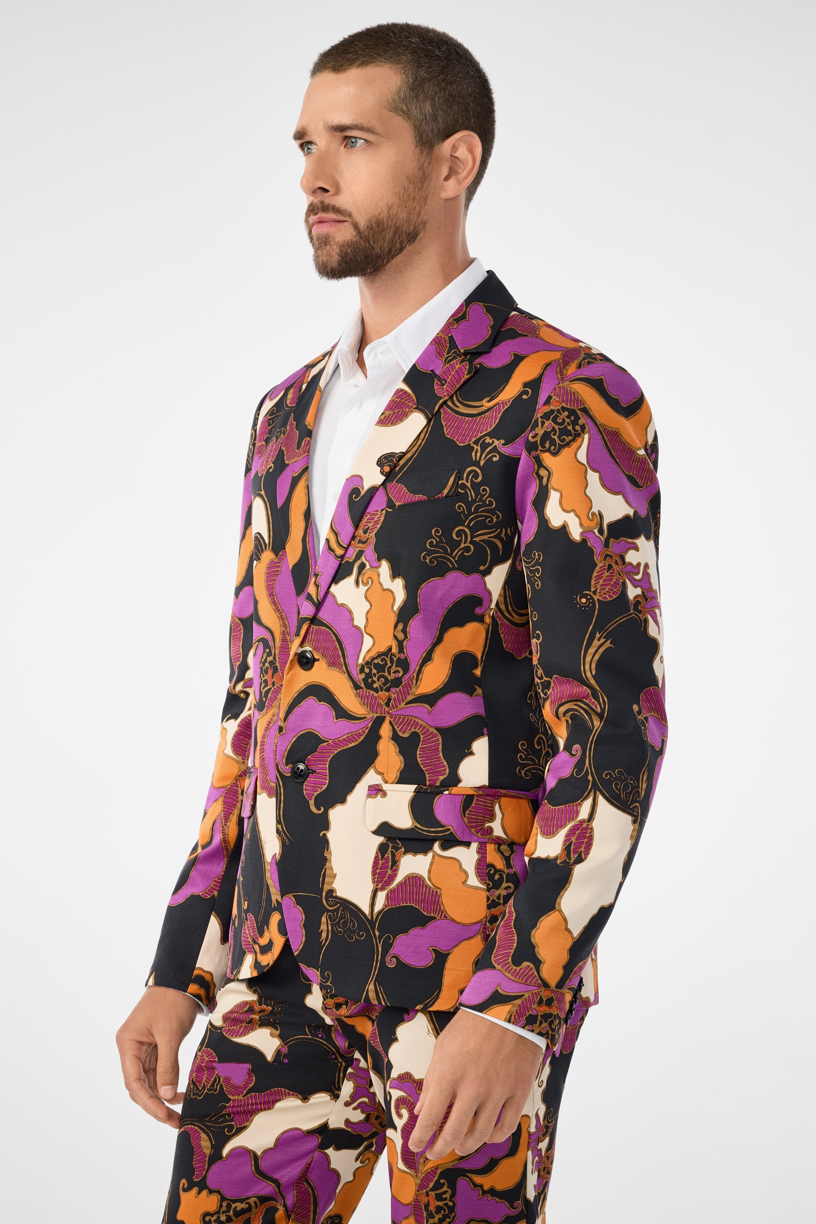A man with a short beard wears the Thurston Blazer, which features an abstract floral pattern in purple, orange, black, and white over a white dress shirt. He stands against a plain white background, looking to the side.