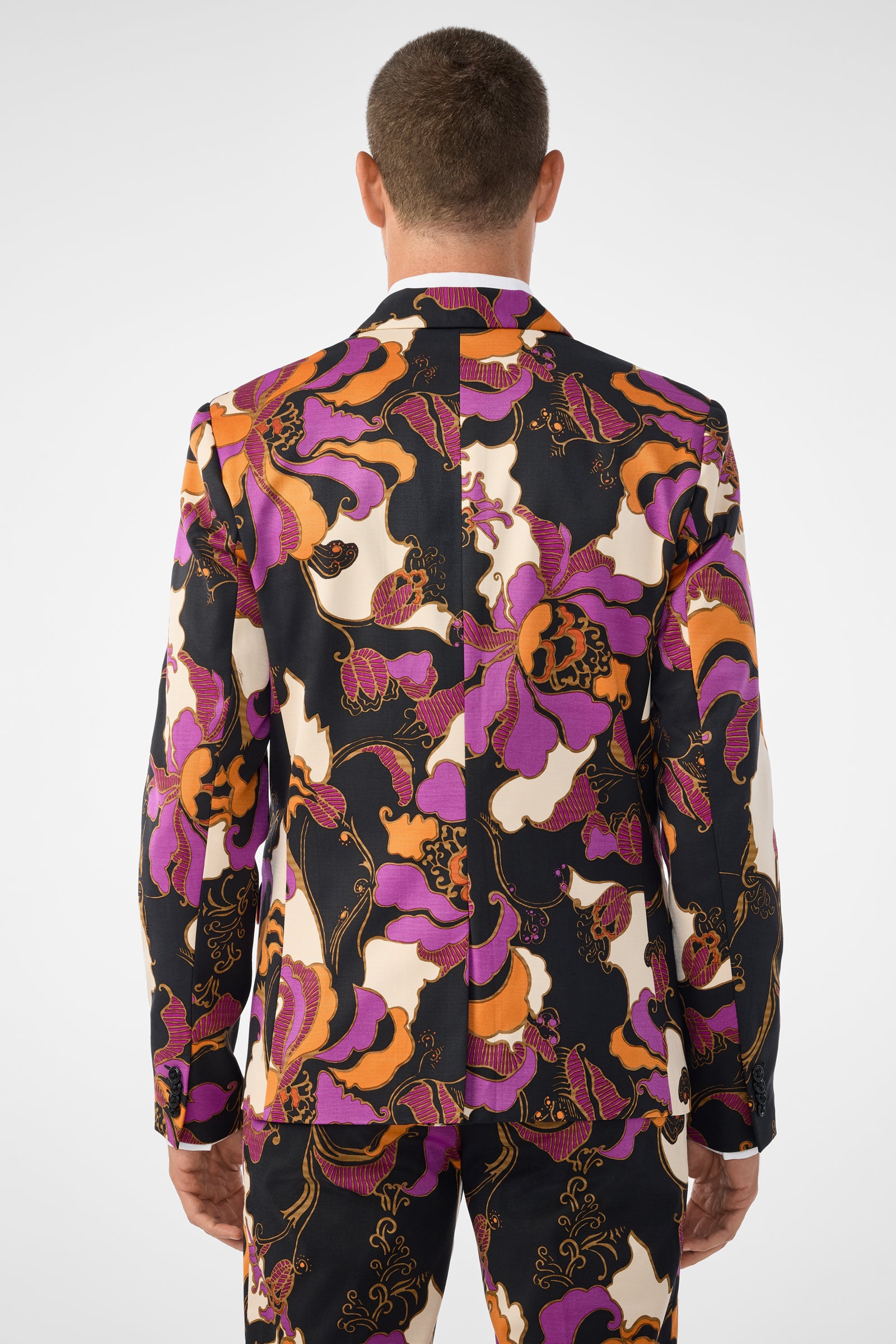 A man is shown from behind wearing the Thurston Blazer, a colorful suit jacket and matching pants with a bold, abstract floral pattern in orange, purple, black, and white. The background is plain white.