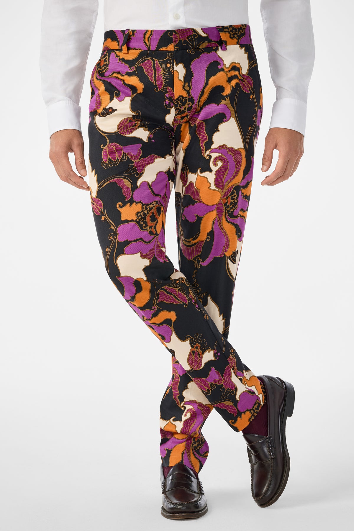A person stands with legs crossed against a plain white background, wearing a white shirt, dark brown dress shoes, and Clyde Slim Trouser featuring a vibrant black, purple, orange, and cream floral pattern.