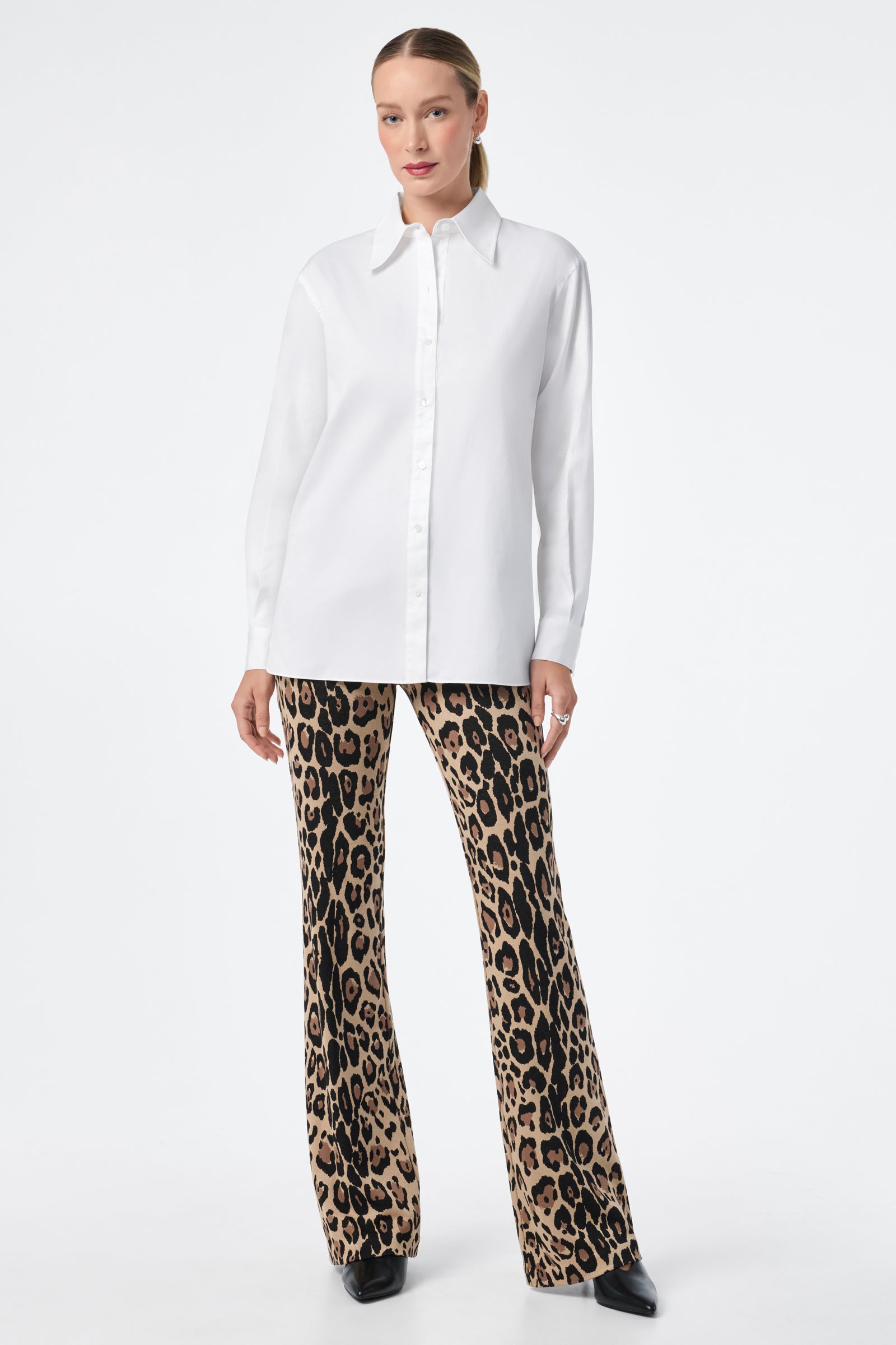 A woman with light hair pulled back and a neutral expression stands against a white background wearing the Litsa Button-Up Cotton Shirt, flared leopard print pants, and black shoes.