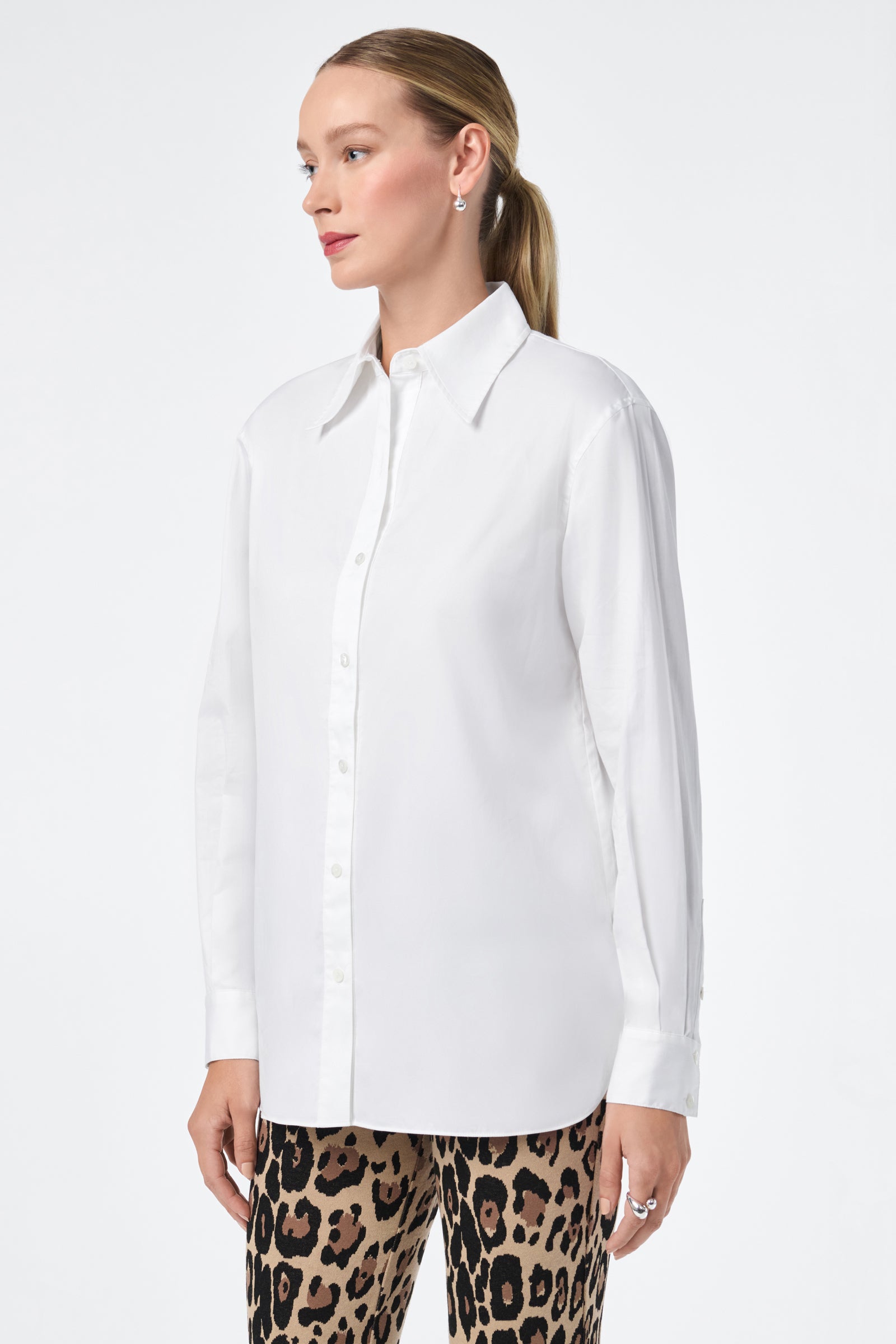 A woman with blonde hair in a ponytail models the Litsa Long-Sleeve Button-Up Cotton Shirt and leopard print pants while standing against a plain white background, facing slightly to the side.