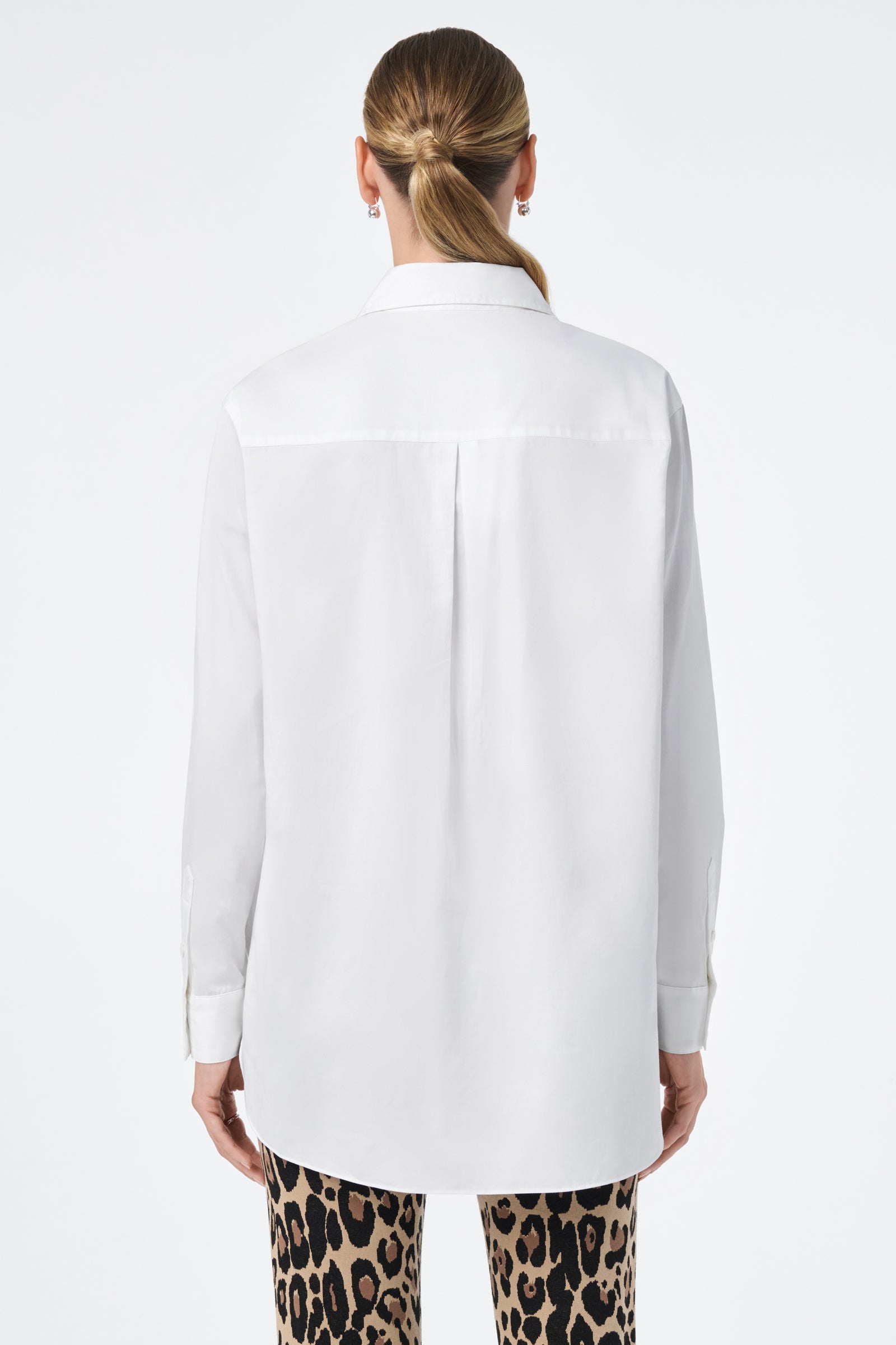 A woman with her hair in a low ponytail faces away from the camera, wearing the Litsa Long-Sleeve Button-Up Cotton Shirt and leopard print pants against a plain white background.