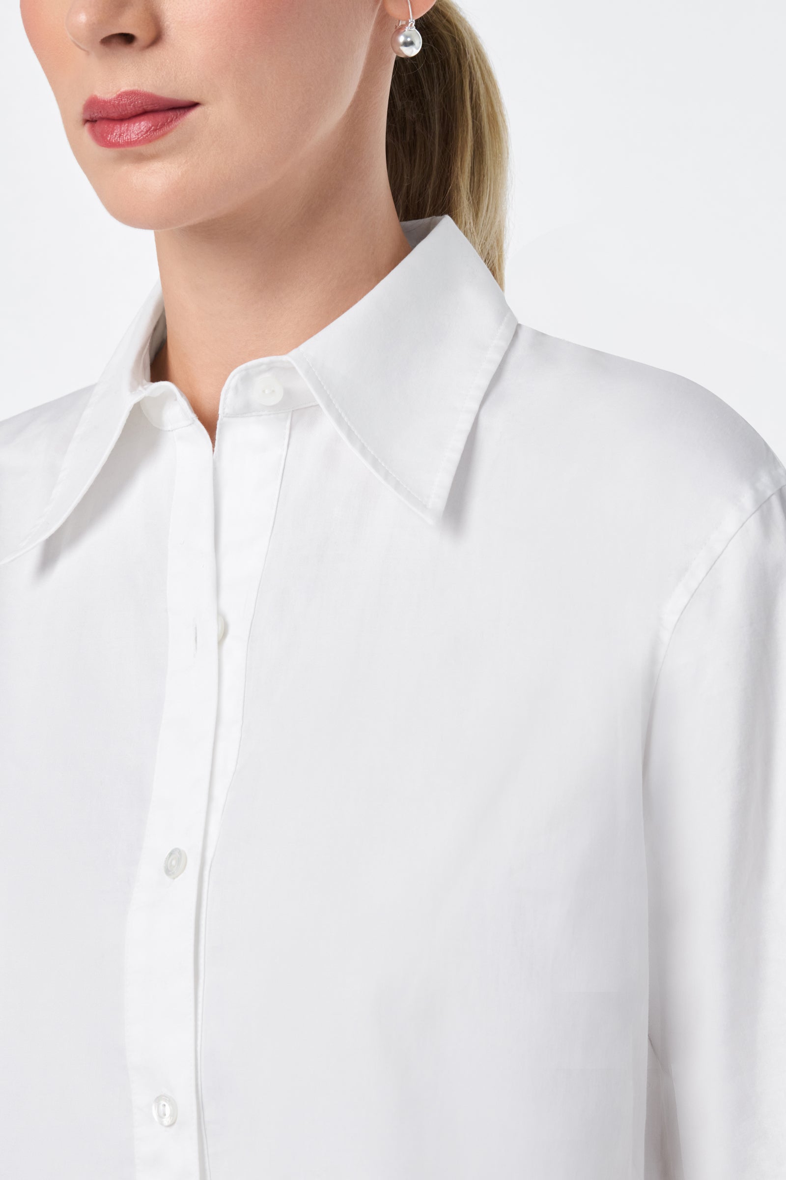 A woman with light skin wears the Litsa Long-Sleeve Button-Up Cotton Shirt with a wide collar. Only the lower half of her face and upper torso are visible, and she accessorizes with a pearl earring.