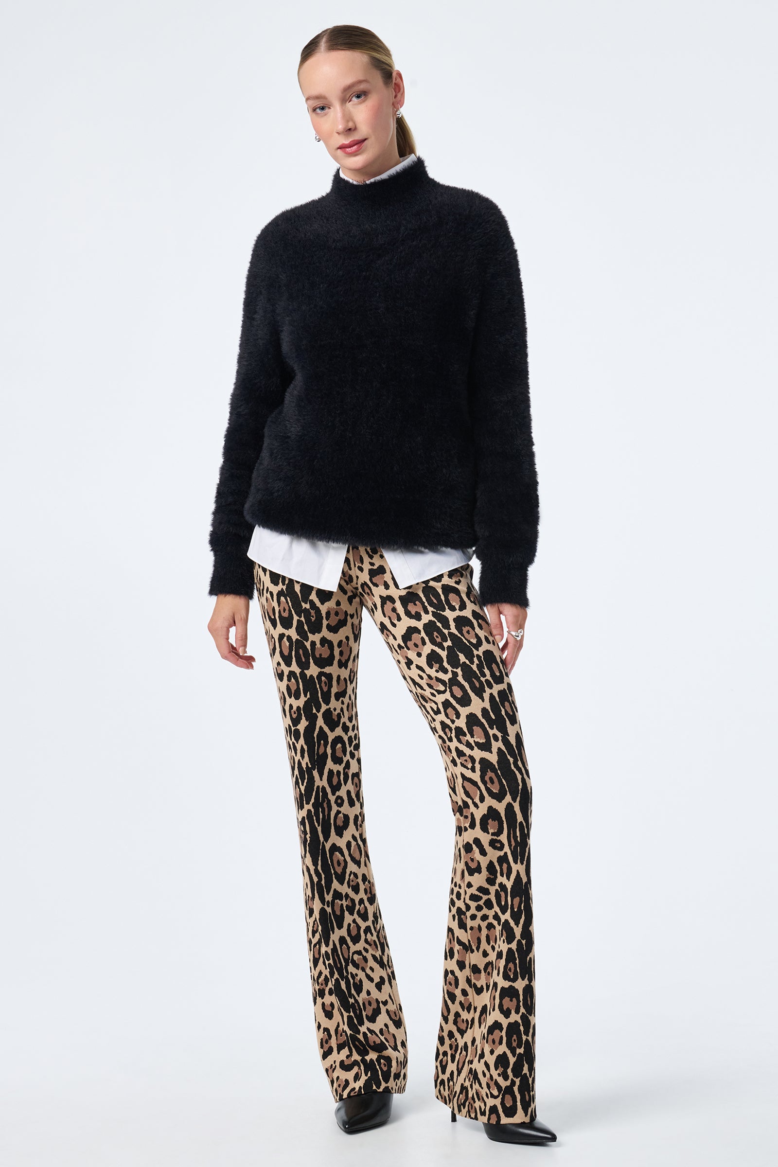 A woman poses casually against a white background wearing the Litsa Button-Up Cotton Shirt under a fuzzy black sweater, paired with high-waisted, flared leopard print pants and black shoes. Her hair is pulled back.