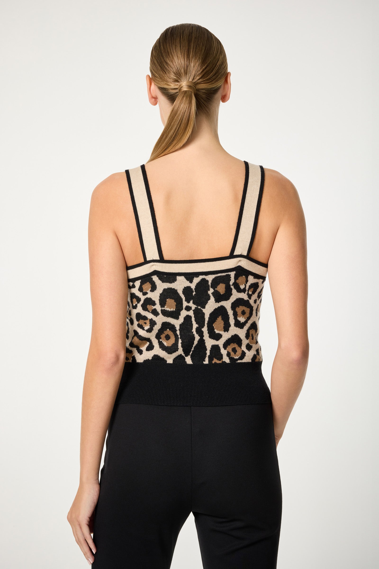 A woman with long, straight hair in a low ponytail is seen from the back wearing the Casady Knit Cami, featuring a beige and black leopard print, paired with black pants against a plain light background.