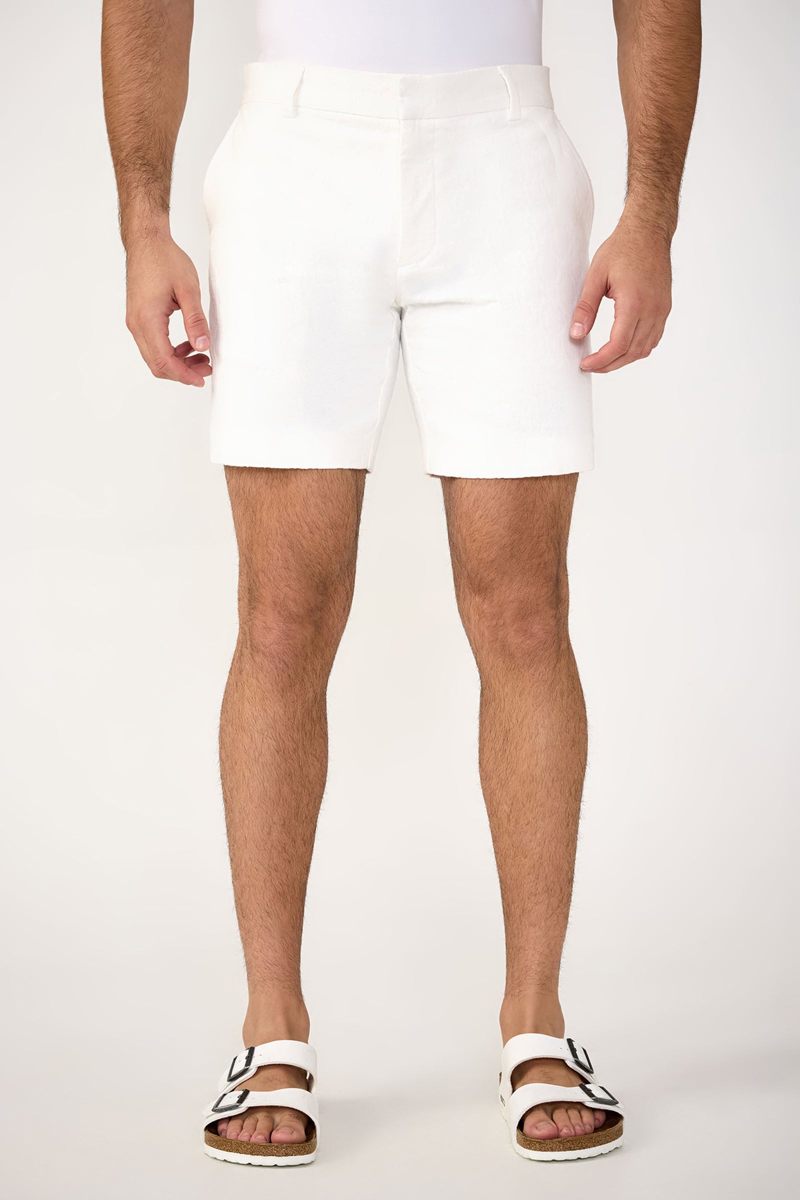 A person wearing the Lawrence Short - Stretch Herringbone in Whitewash stands against a plain light background, paired with white buckled sandals. Only the lower torso and legs are visible.