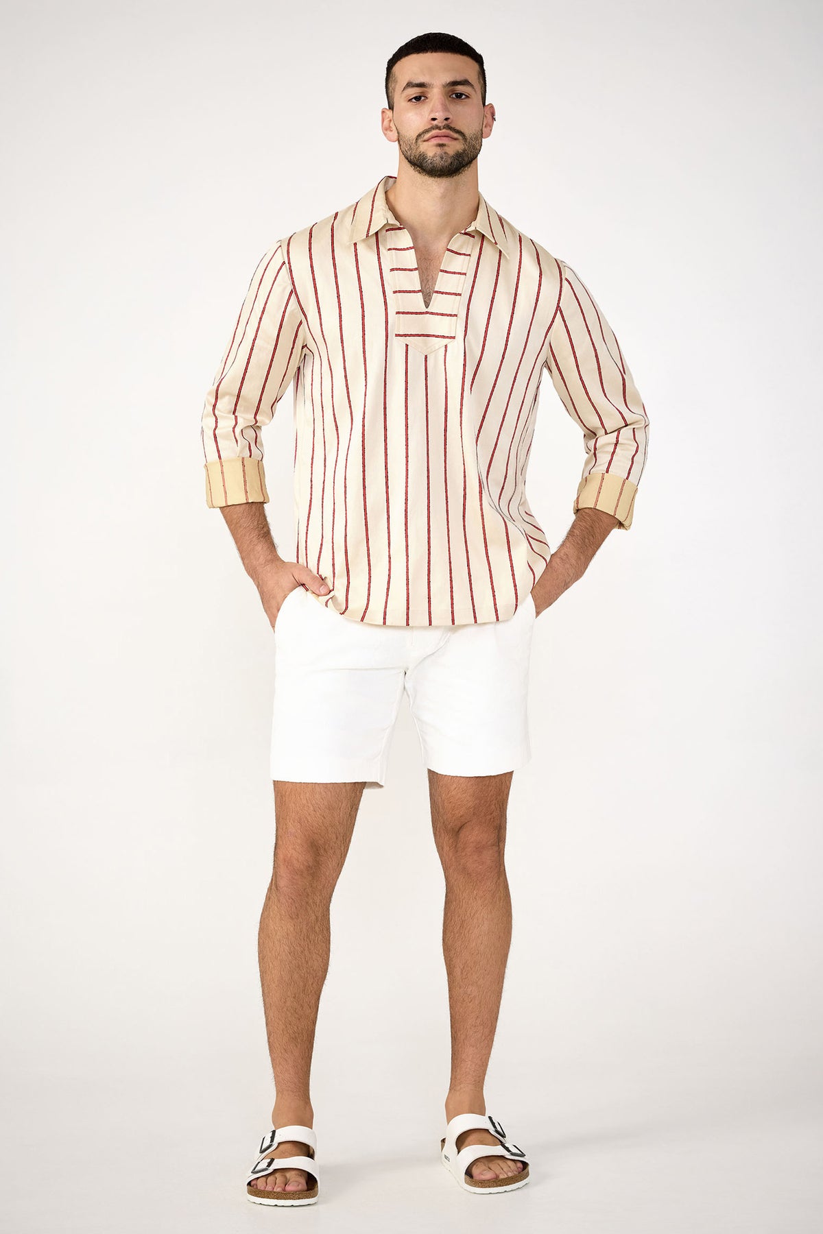 A man with a short beard poses against a plain background, wearing the Lawrence Short - Stretch Herringbone in white, paired with a cream shirt featuring vertical red stripes and white slide sandals. His hands are in his pockets as he faces forward.