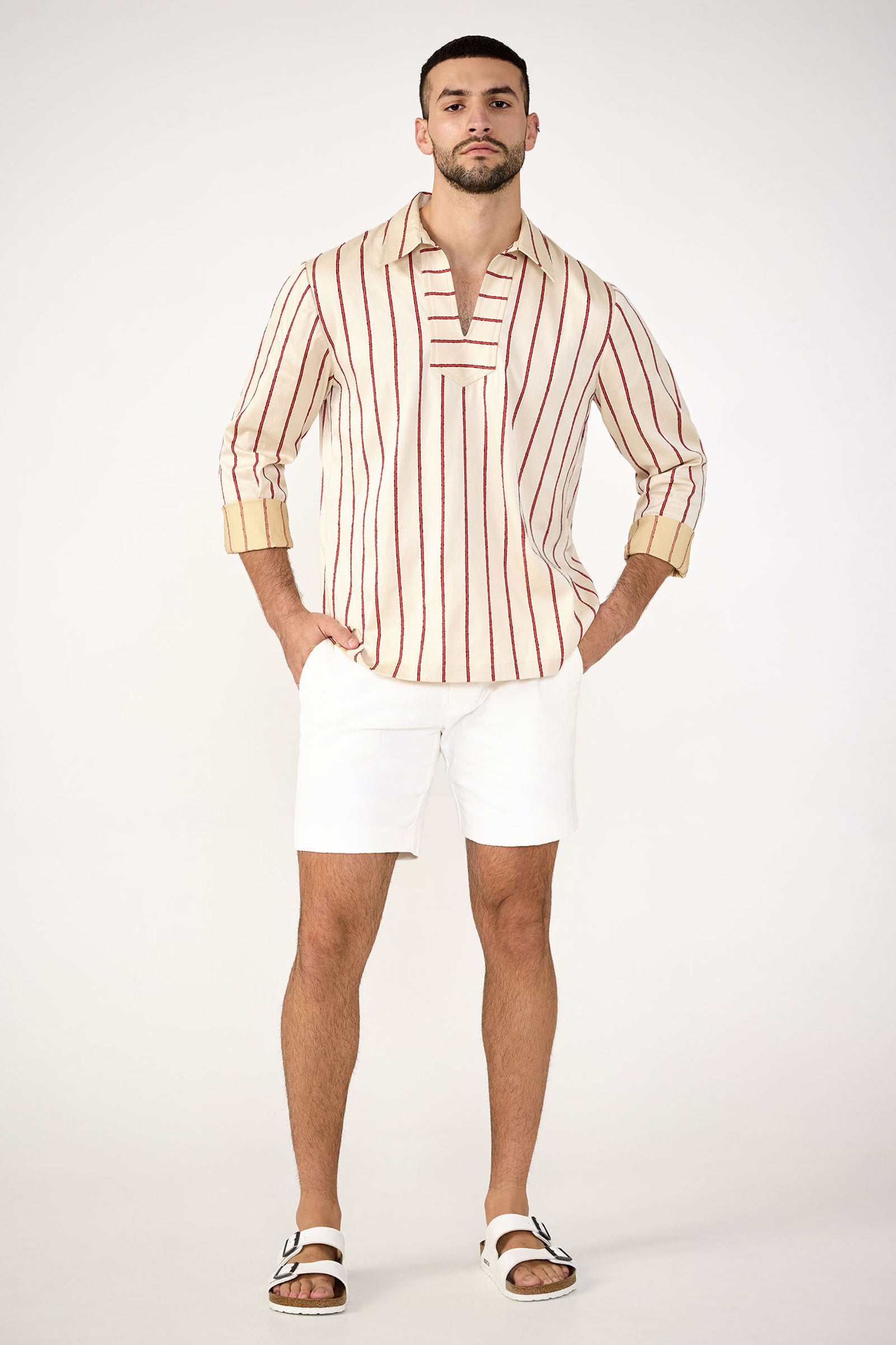 A man with a short beard poses against a plain background, wearing the Lawrence Short - Stretch Herringbone in white, paired with a cream shirt featuring vertical red stripes and white slide sandals. His hands are in his pockets as he faces forward.
