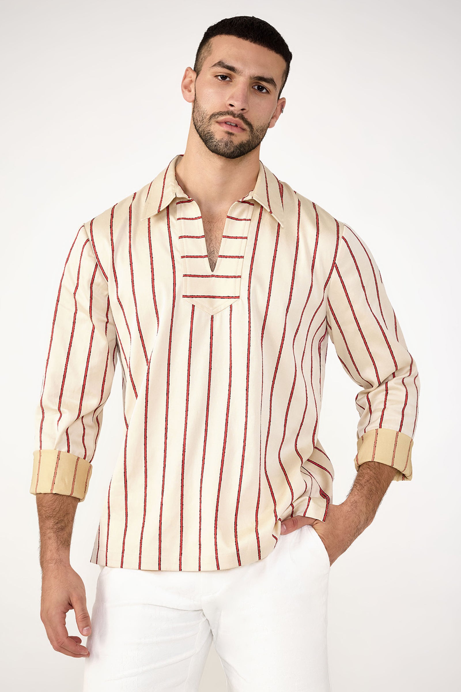 A man confidently poses against a plain light background, wearing the Holmes Long-Sleeve Pullover in Solaire Stripe with vertical red stripes and beige cuffs, paired with white pants.
