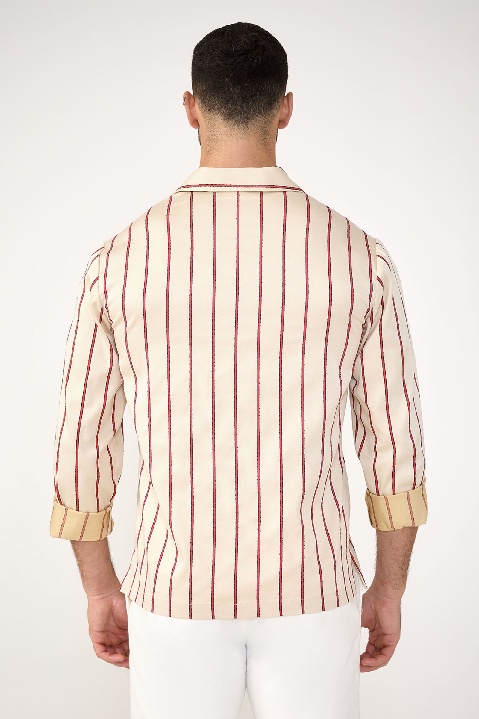 A man faces away from the camera, wearing the Holmes Long-Sleeve Pullover in Solaire Stripe—cream with vertical red and navy stripes and rolled-up sleeves—paired with white pants against a plain light background.