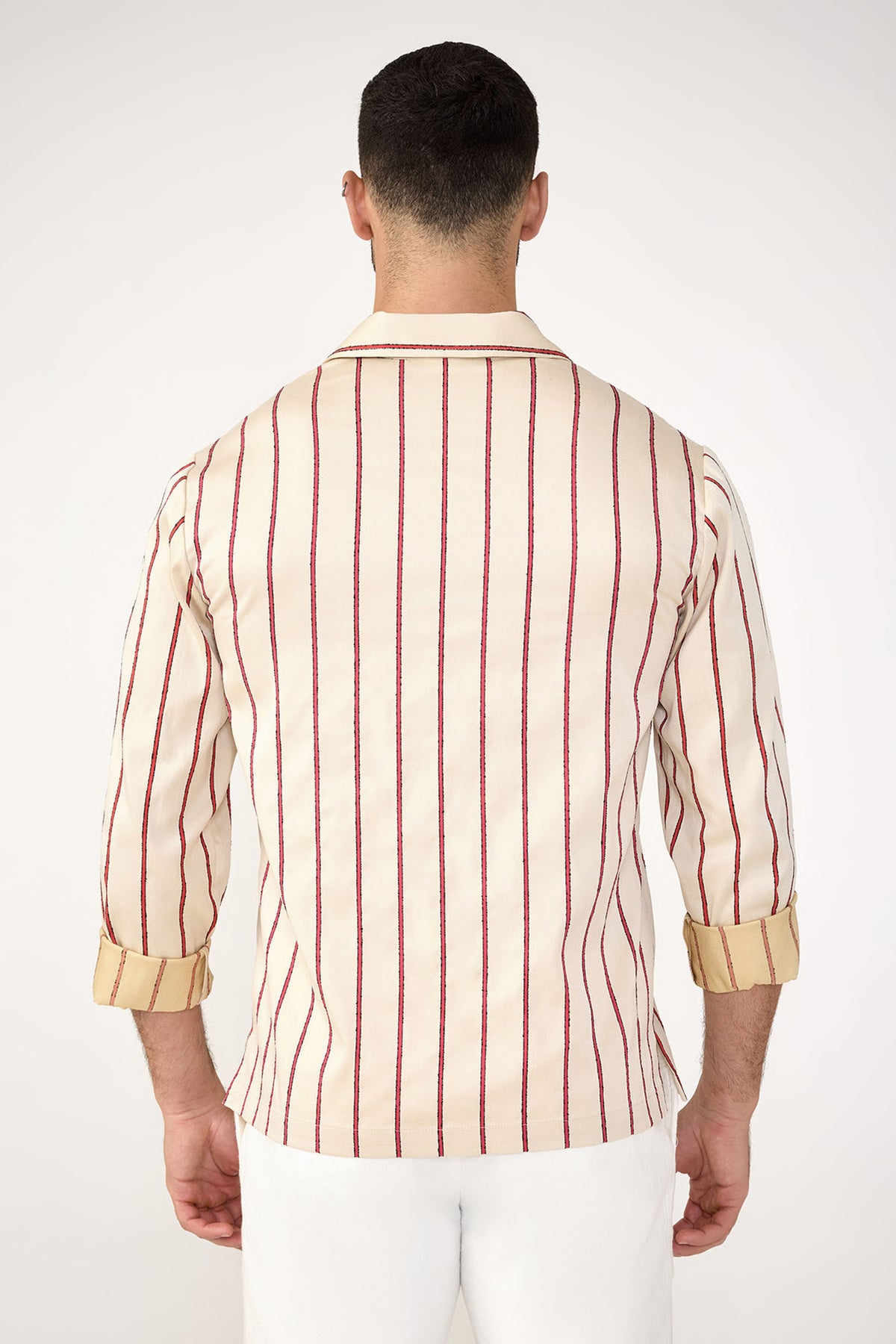 A man faces away from the camera, wearing the Holmes Long-Sleeve Pullover in Solaire Stripe—cream with vertical red and navy stripes and rolled-up sleeves—paired with white pants against a plain light background.
