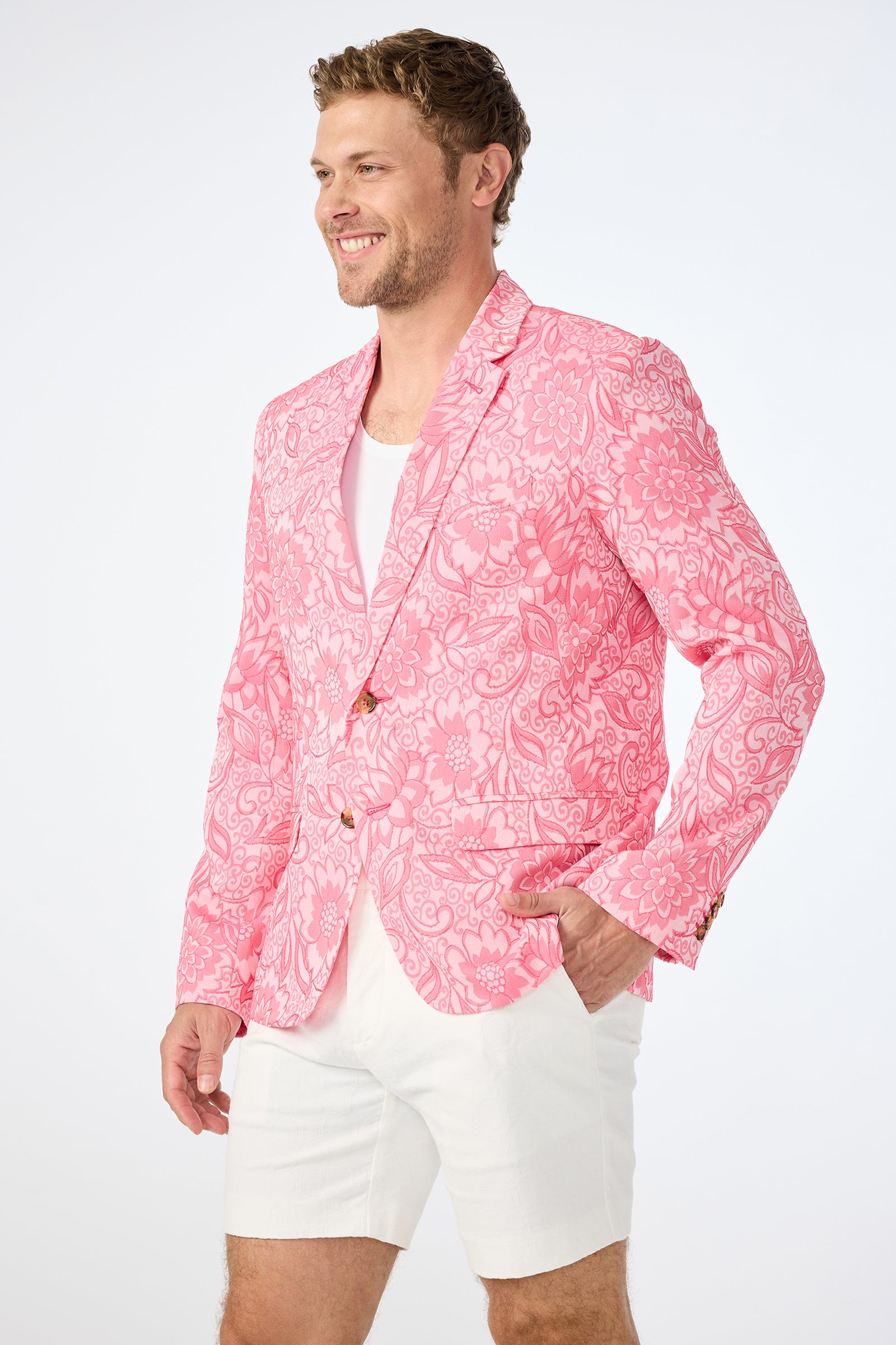 A man wearing the Thurston Blazer - Jolie Jacquard, a bright pink floral-patterned jacket, over a white shirt and white shorts smiles against a plain white background.