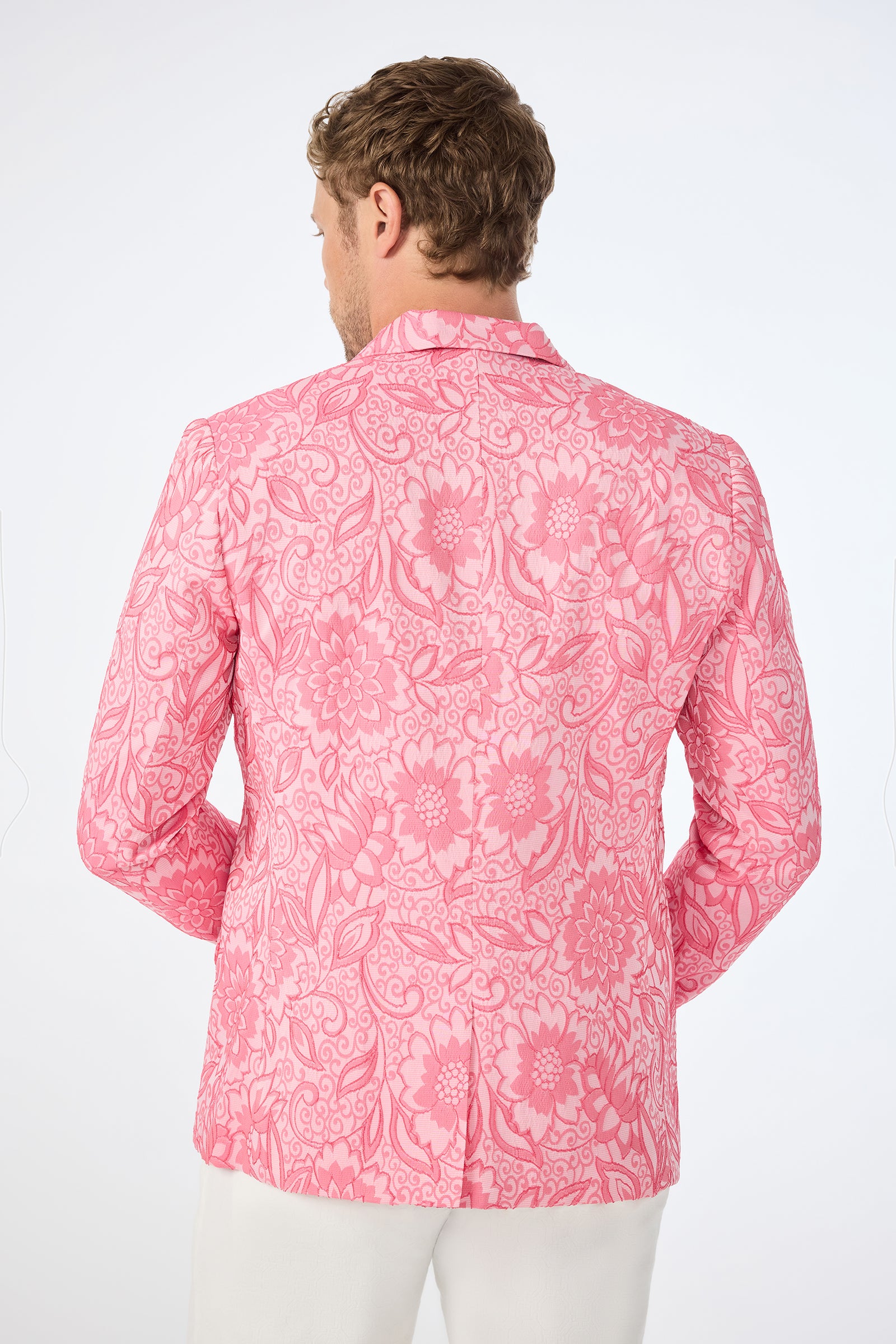 A man with light brown hair is seen from behind, wearing the Thurston Blazer - Jolie Jacquard, featuring an intricate bright pink floral and paisley pattern. He pairs it with white pants and stands against a plain white background.
