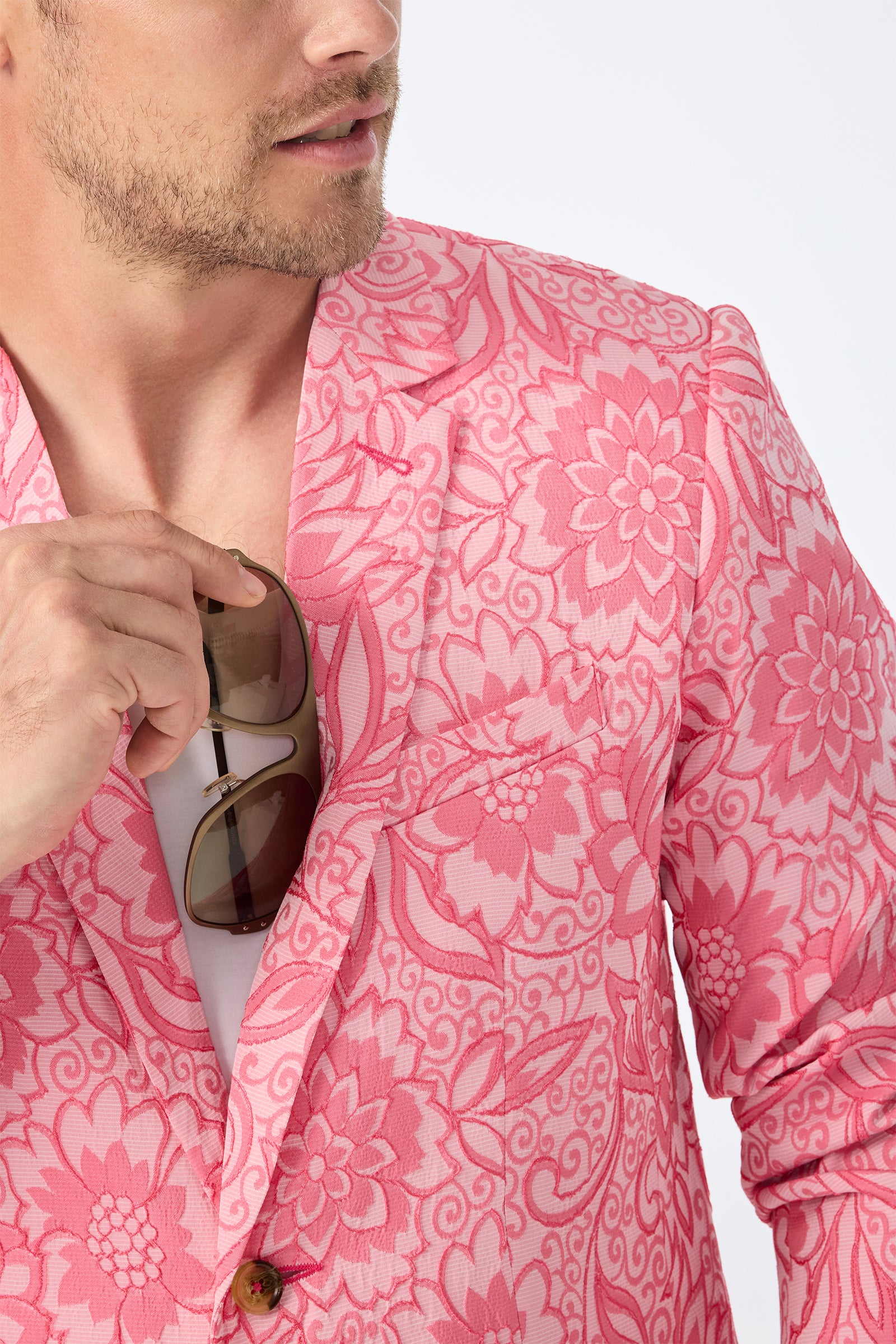A man in the Thurston Blazer - Jolie Jacquard with a pink floral pattern holds sunglasses to his chest. He wears a casual white shirt underneath, and his face is partially visible.