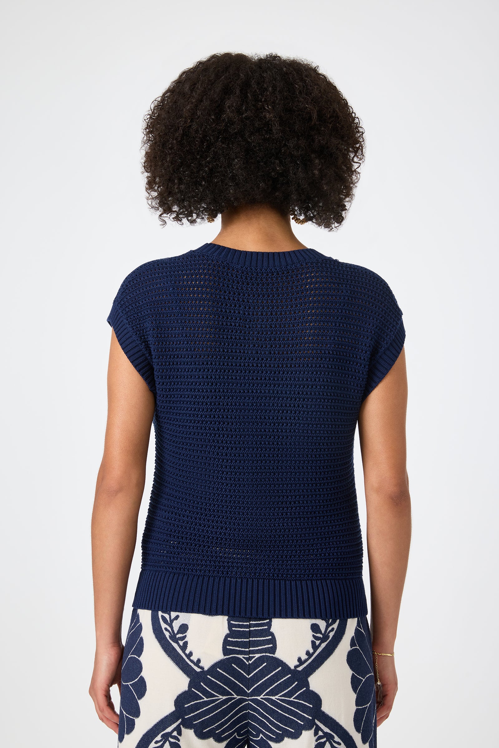 A person with curly hair is shown from behind, wearing the Yuri Knit Pullover in dark blue and a white skirt with a bold navy floral print. The background is plain and light-colored.