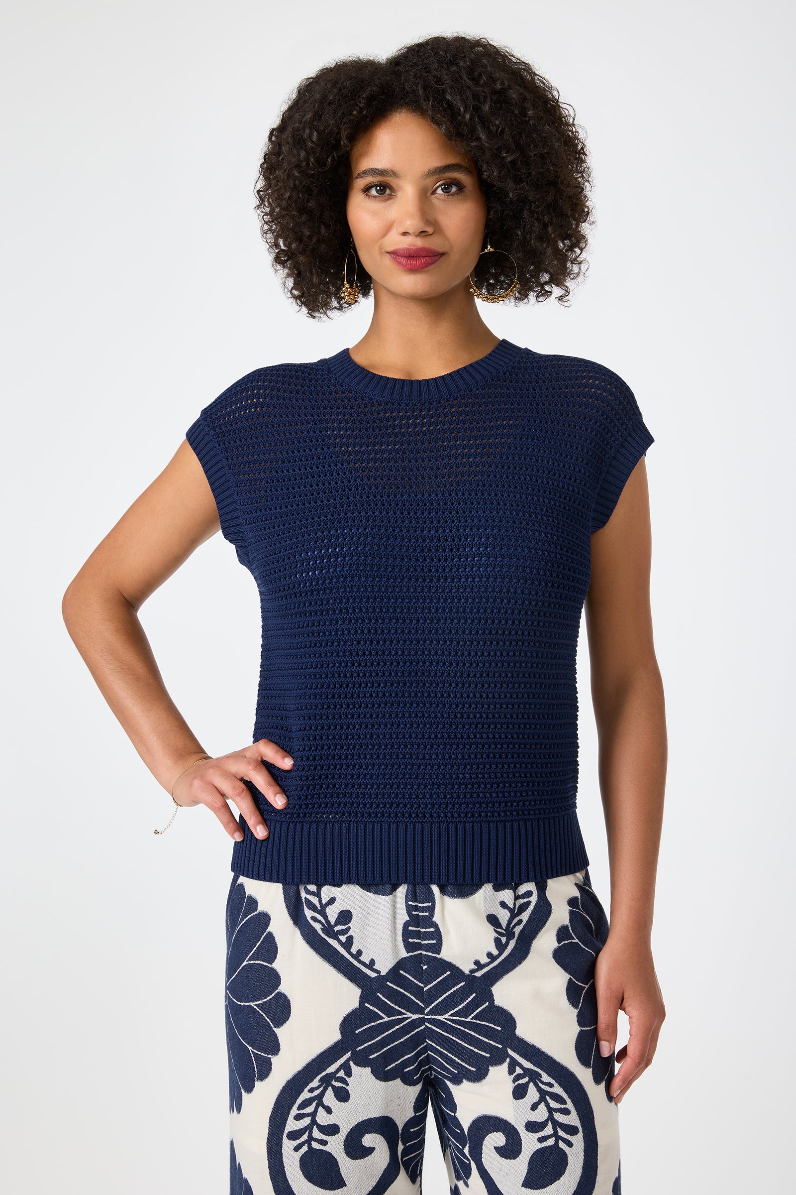 A woman with curly hair wears the Yuri Knit Pullover in Sumi Blue and white pants with a bold navy floral pattern. She stands smiling, one hand on her hip, against a plain background.