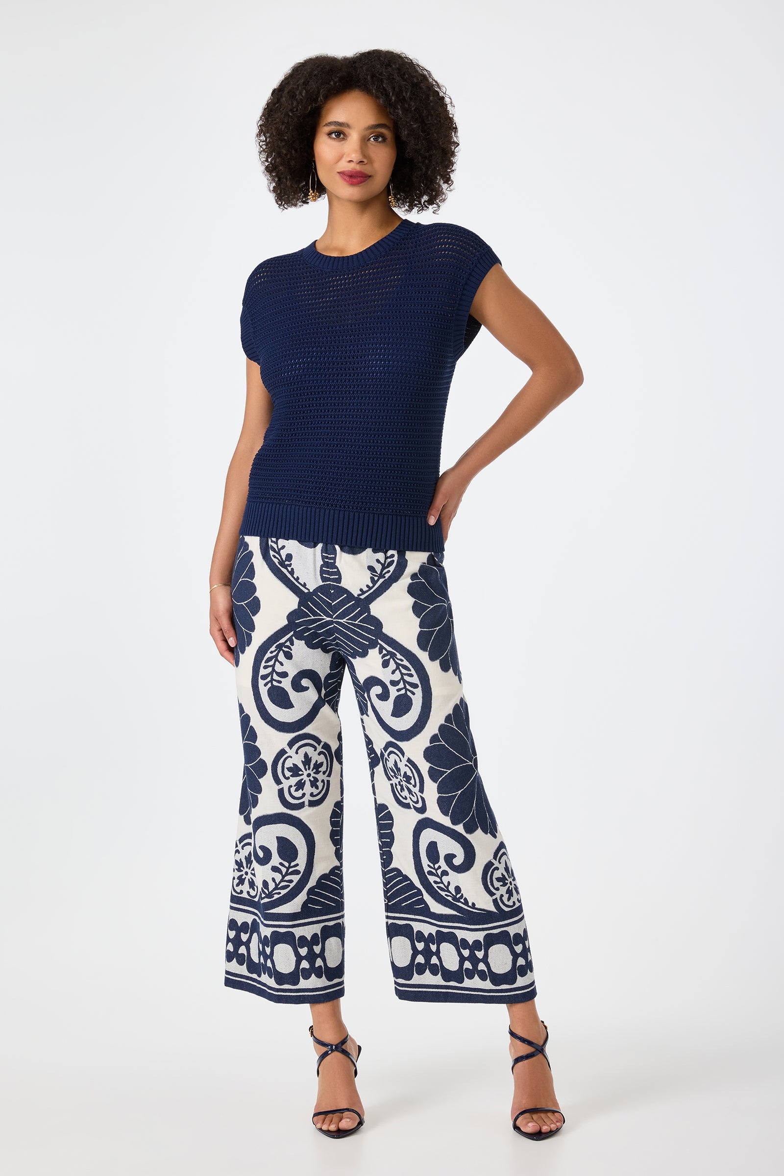 A woman with curly hair confidently poses against a white background wearing the Yuri Knit Pullover, wide-leg white pants with a bold navy floral pattern, and black strappy heels.