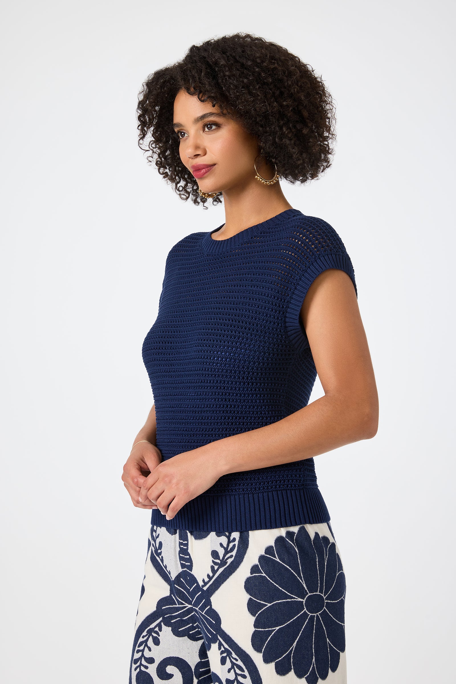 A woman with curly hair models the Yuri Knit Pullover in navy blue, paired with a white skirt featuring bold blue floral and vine patterns, against a plain light background.