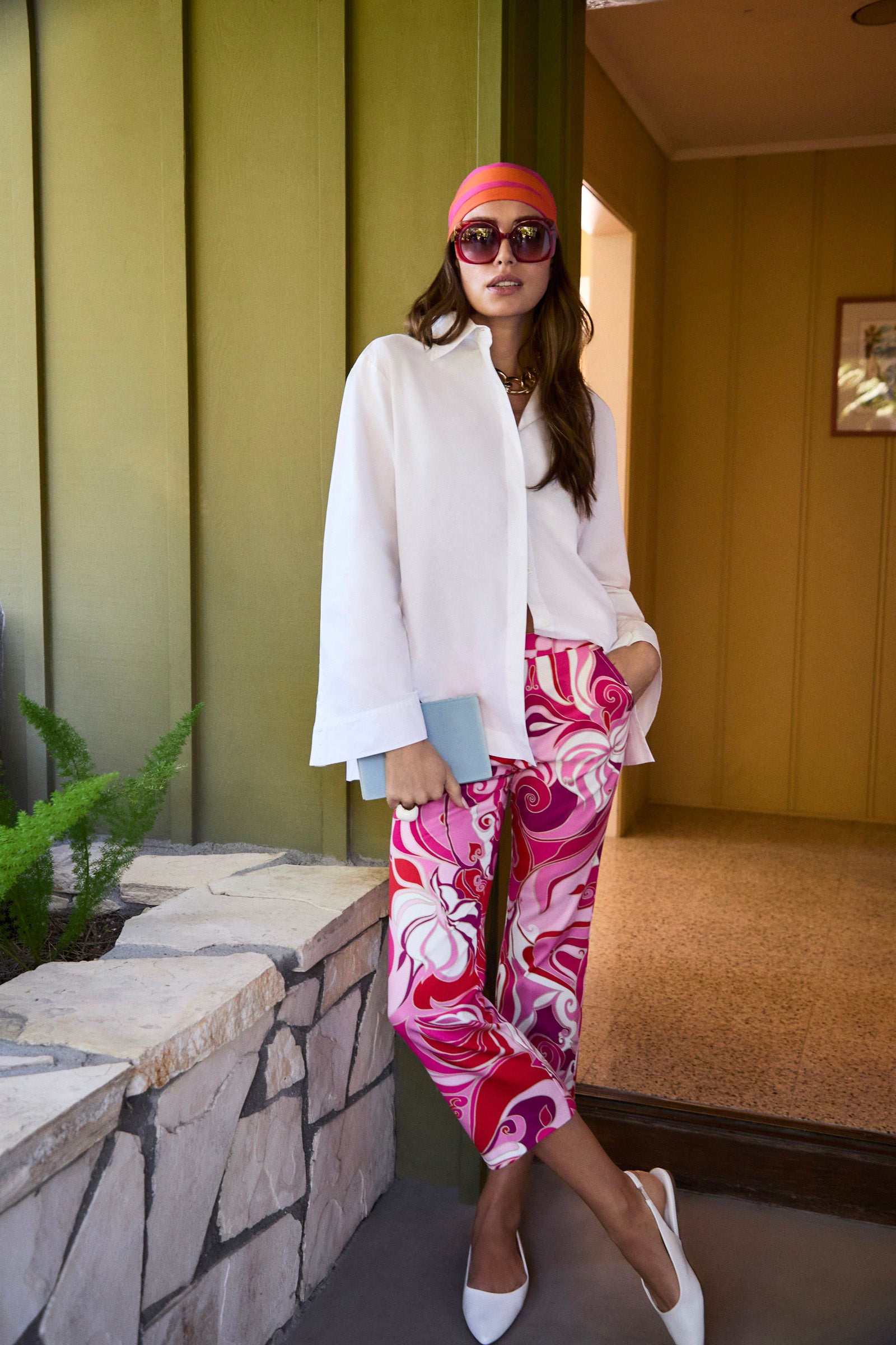 Wearing the Olona Swing Top, a woman pairs it with vibrant pink patterned pants by a stone ledge. She completes the look with a coral headscarf, sunglasses, white heels, and a light blue clutch.